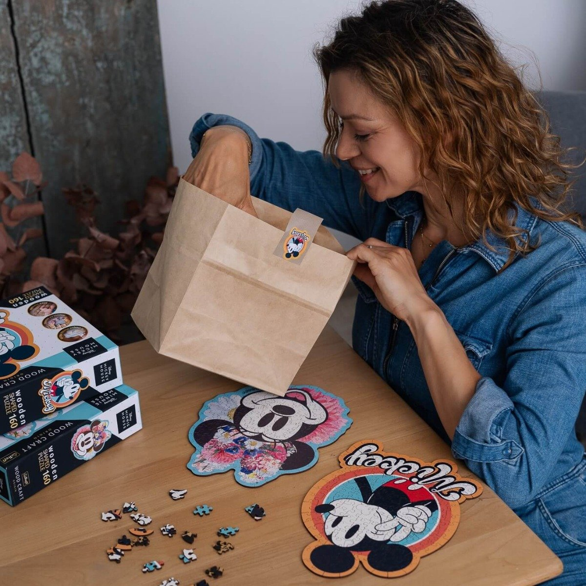 A woman opening a brown paper bag, with Minnie Mouse puzzles and puzzle pieces on a wooden table.