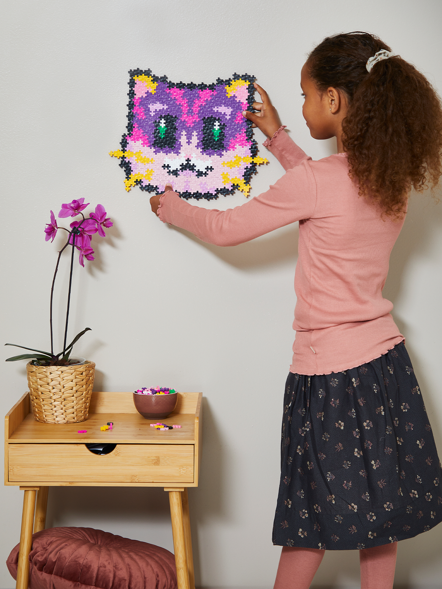 Dziewczyna wiesza na ścianie gotową układankę z kotkiem. Jest stół z orchideą i kilkoma elementami.