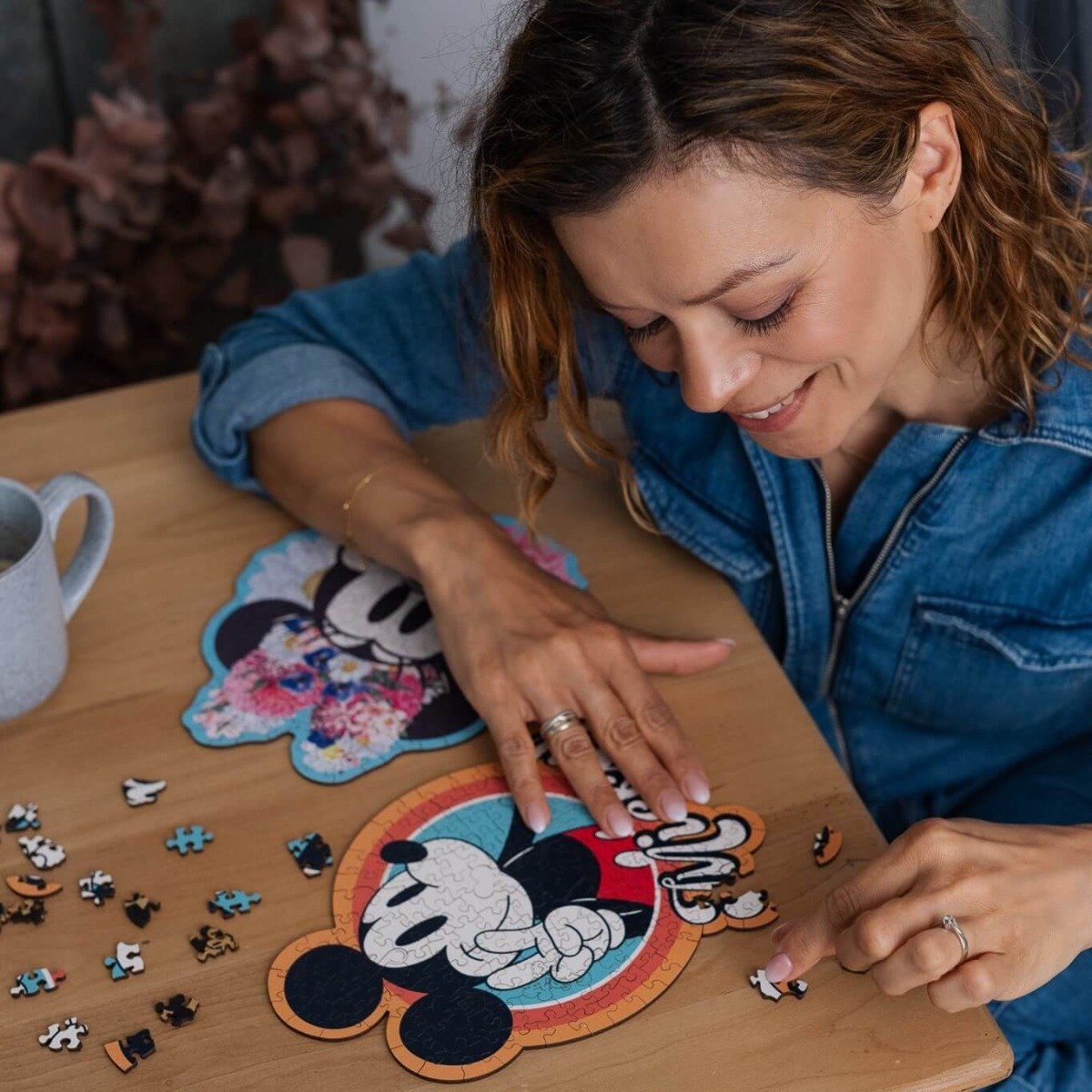 Woman assembling a Mickey Mouse puzzle on a wooden table. Other puzzle pieces are scattered around.