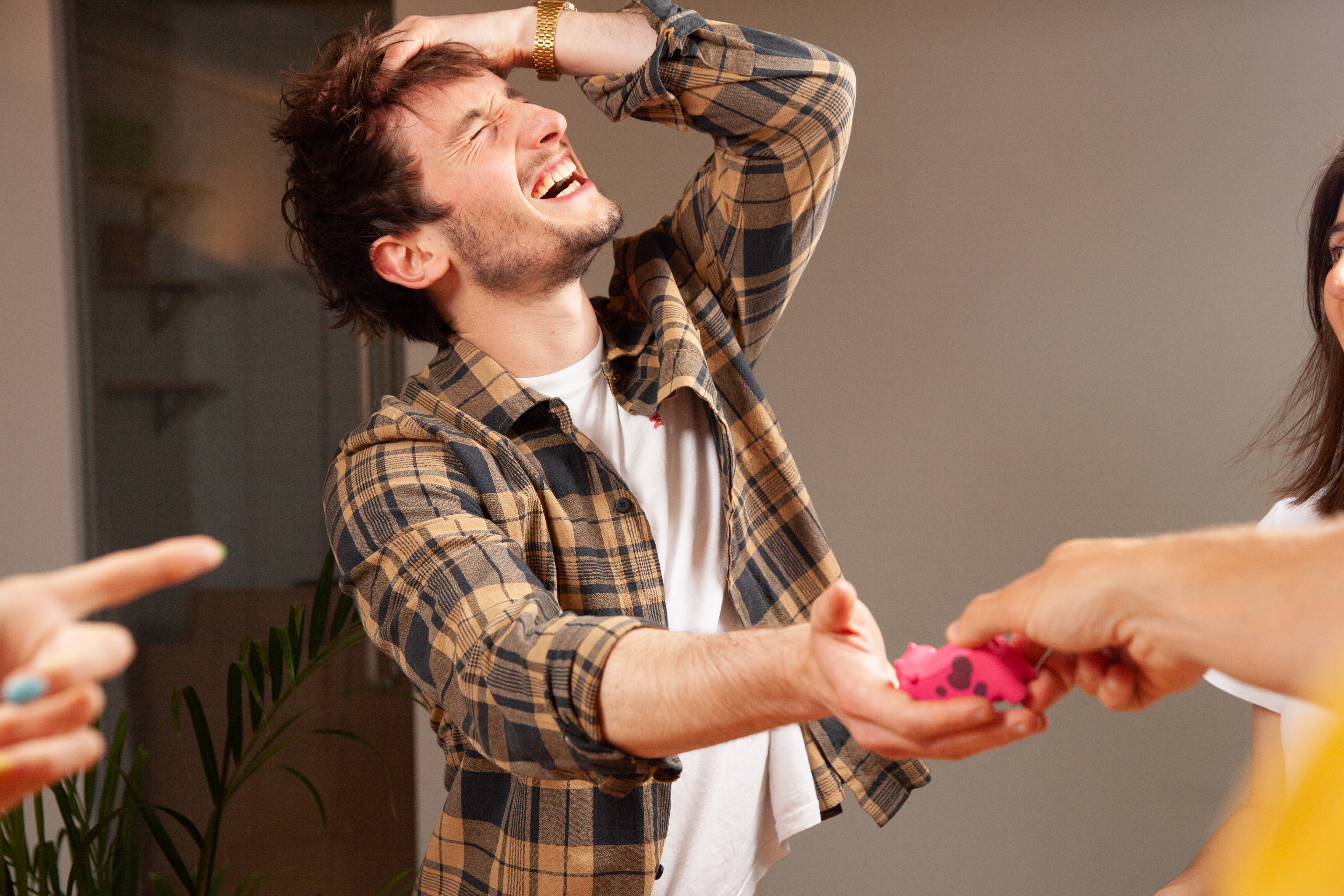 Ein Mann lacht, während er ein rosa Spielzeug hält; zwei weitere Personen sind im Hintergrund.