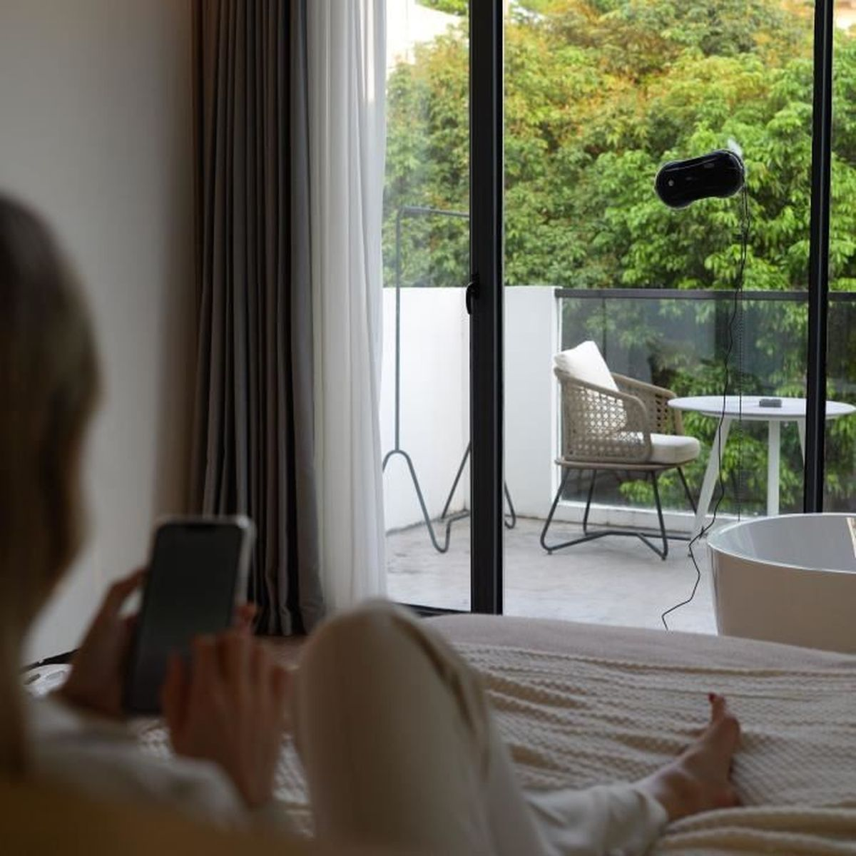 Kobieta na łóżku używa telefonu, patrząc na balkon z robotem do czyszczenia okien na szkle.