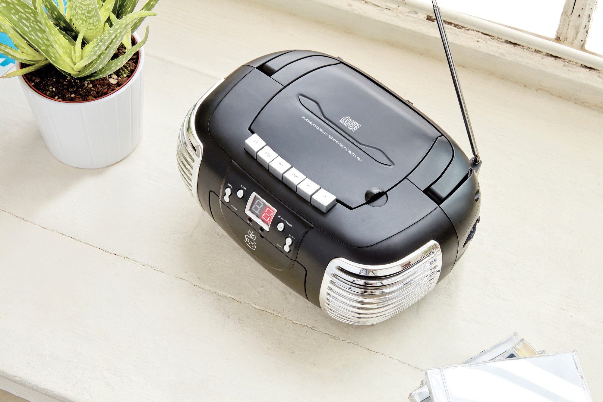 Czarny boombox ze srebrnymi akcentami, odtwarzaczem CD i wyświetlaczem cyfrowym. W scenie roślina i płyty CD.