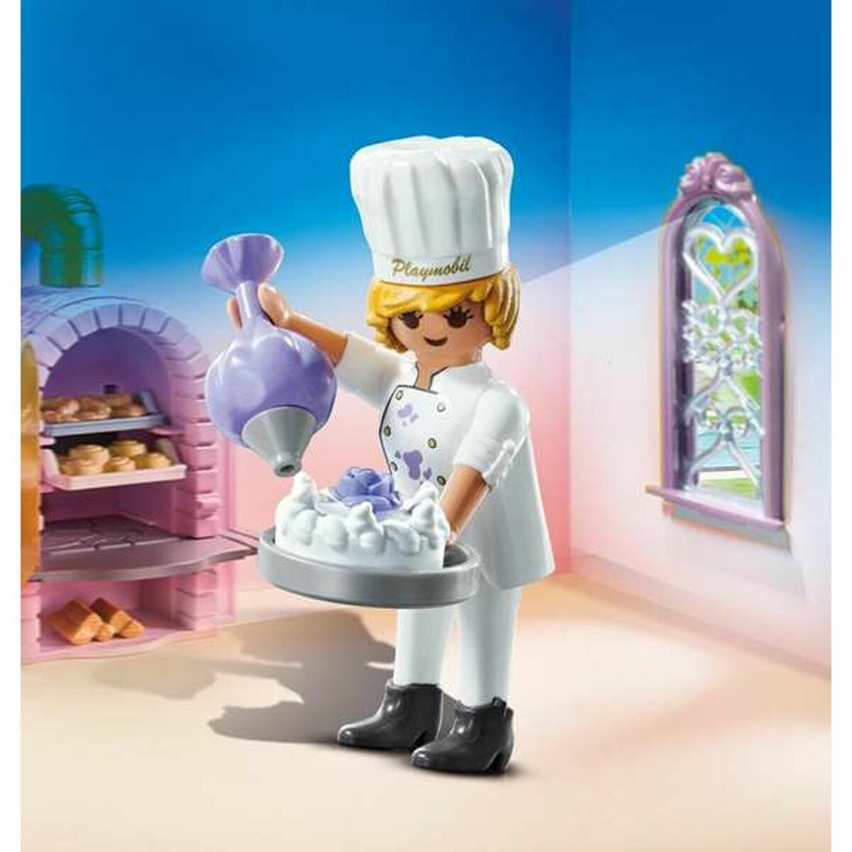 Un cuoco Playmobil che decora una torta con la glassa. Scena della cucina con forno e finestra sullo sfondo.