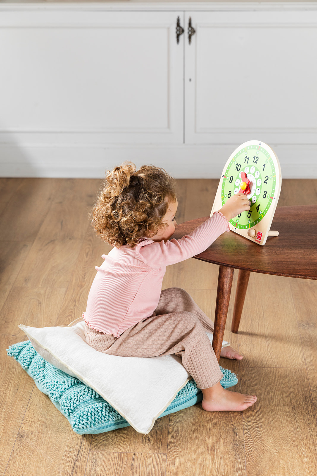 Dziecko siedzi na poduszkach, wchodząc w interakcję z zegarem na stole. Tło: biała szafka, drewniana podłoga.