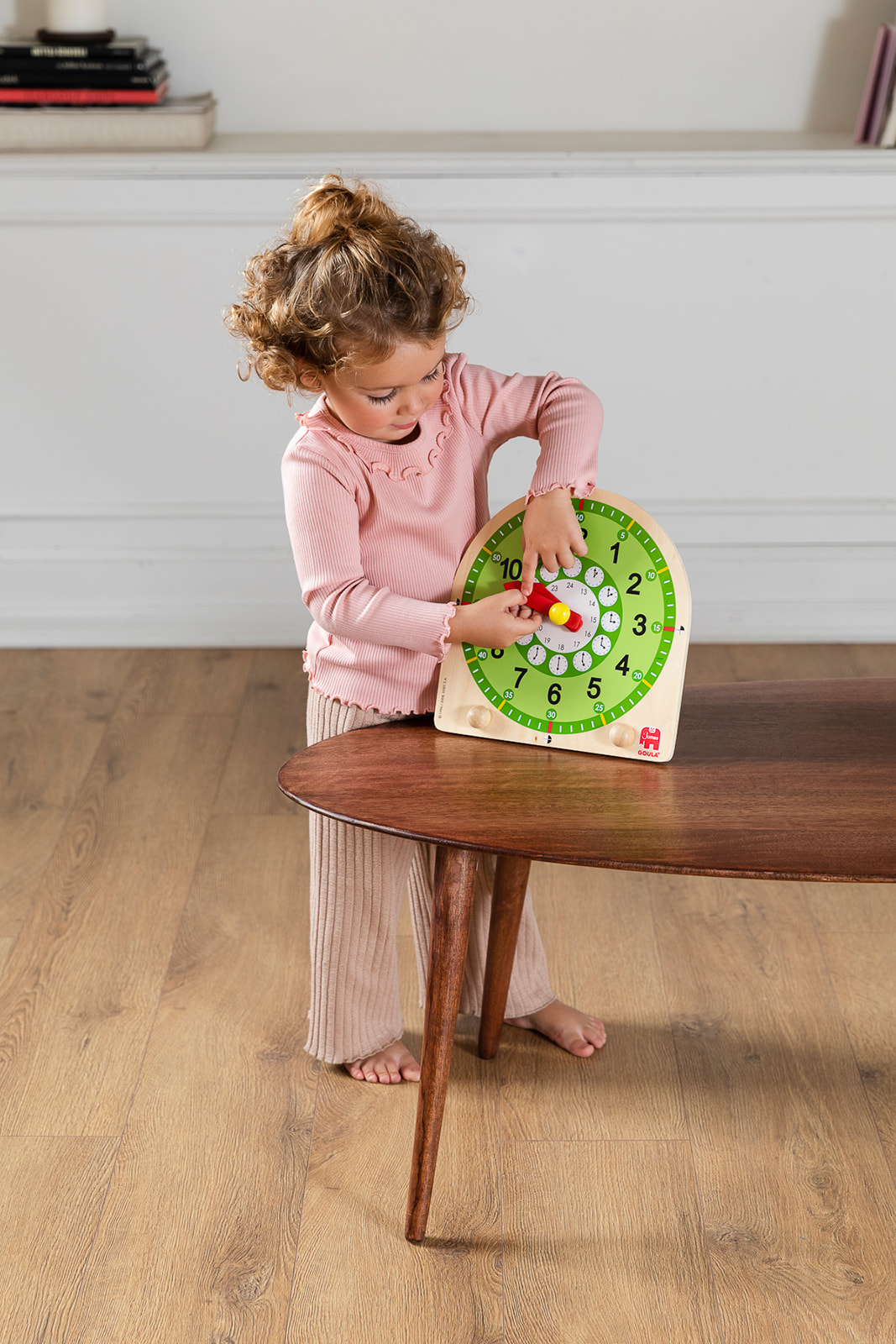 Dziecko wchodzi w interakcję z zegarem na stole. Tło stanowi drewniana podłoga.