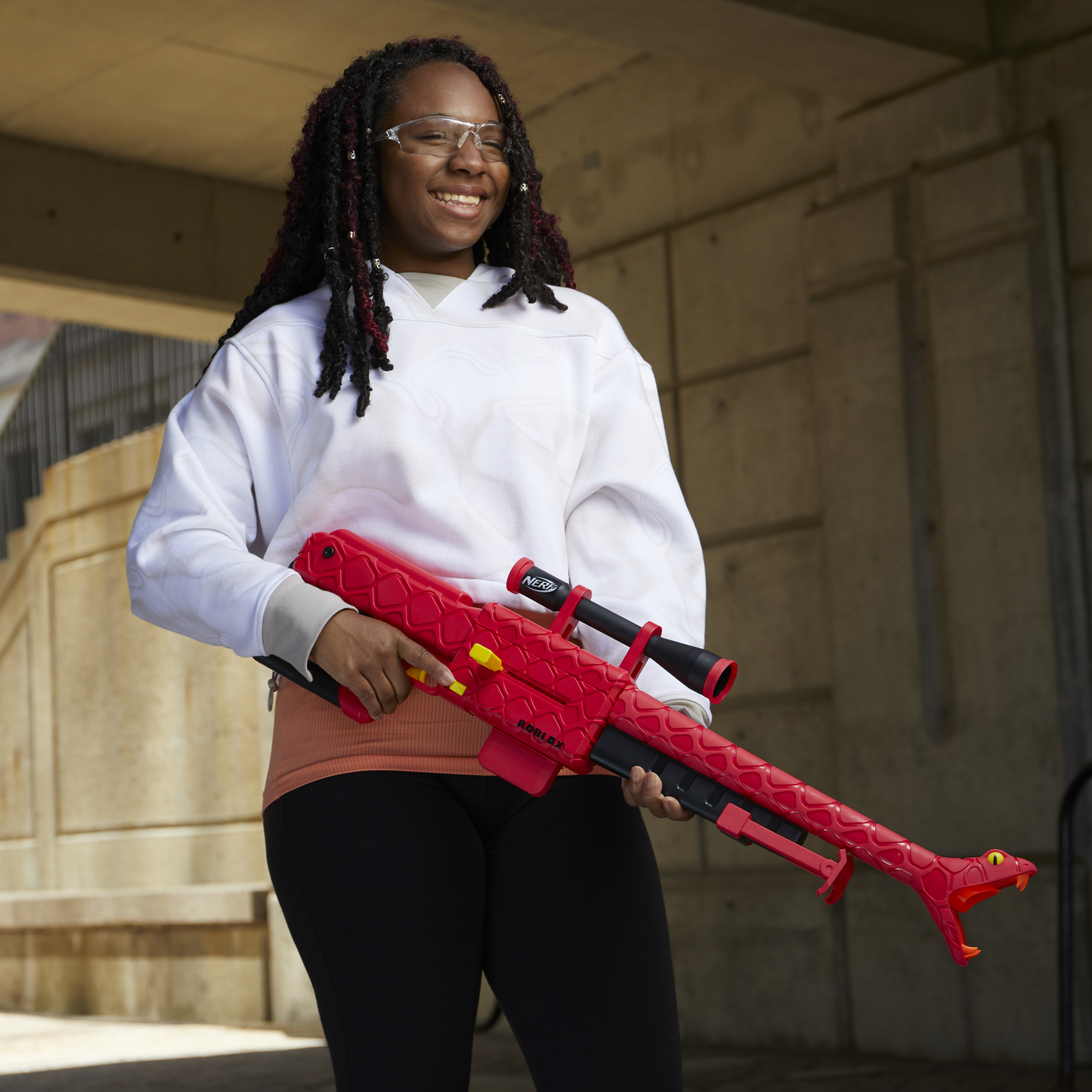 Kobieta trzymająca czerwony karabin Nerf Roblox, uśmiechnięta. W tle budynek.