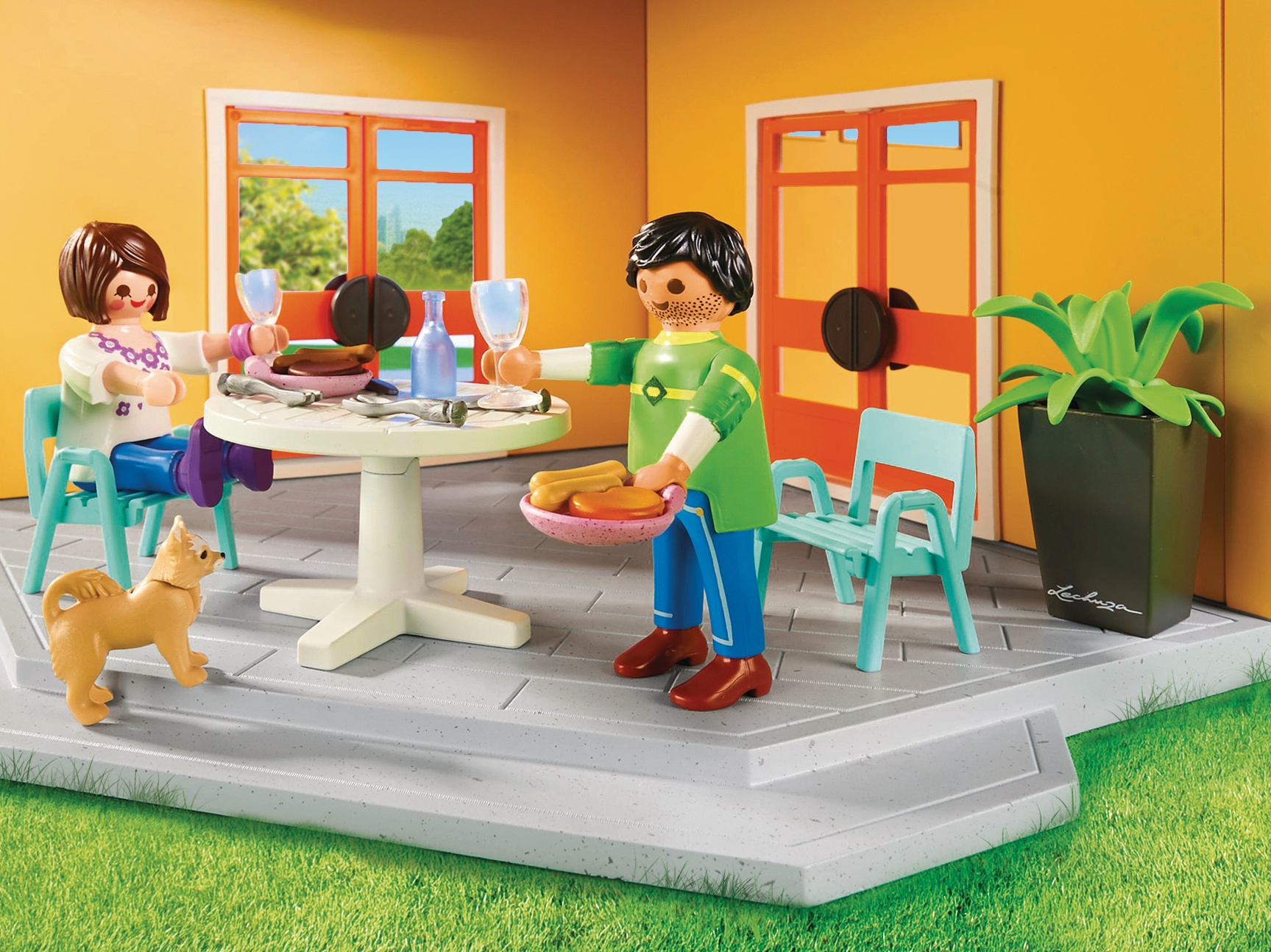 Figurki Playmobil przy stole z jedzeniem i napojami. W pobliżu pies. Tło: dom i zielona trawa.