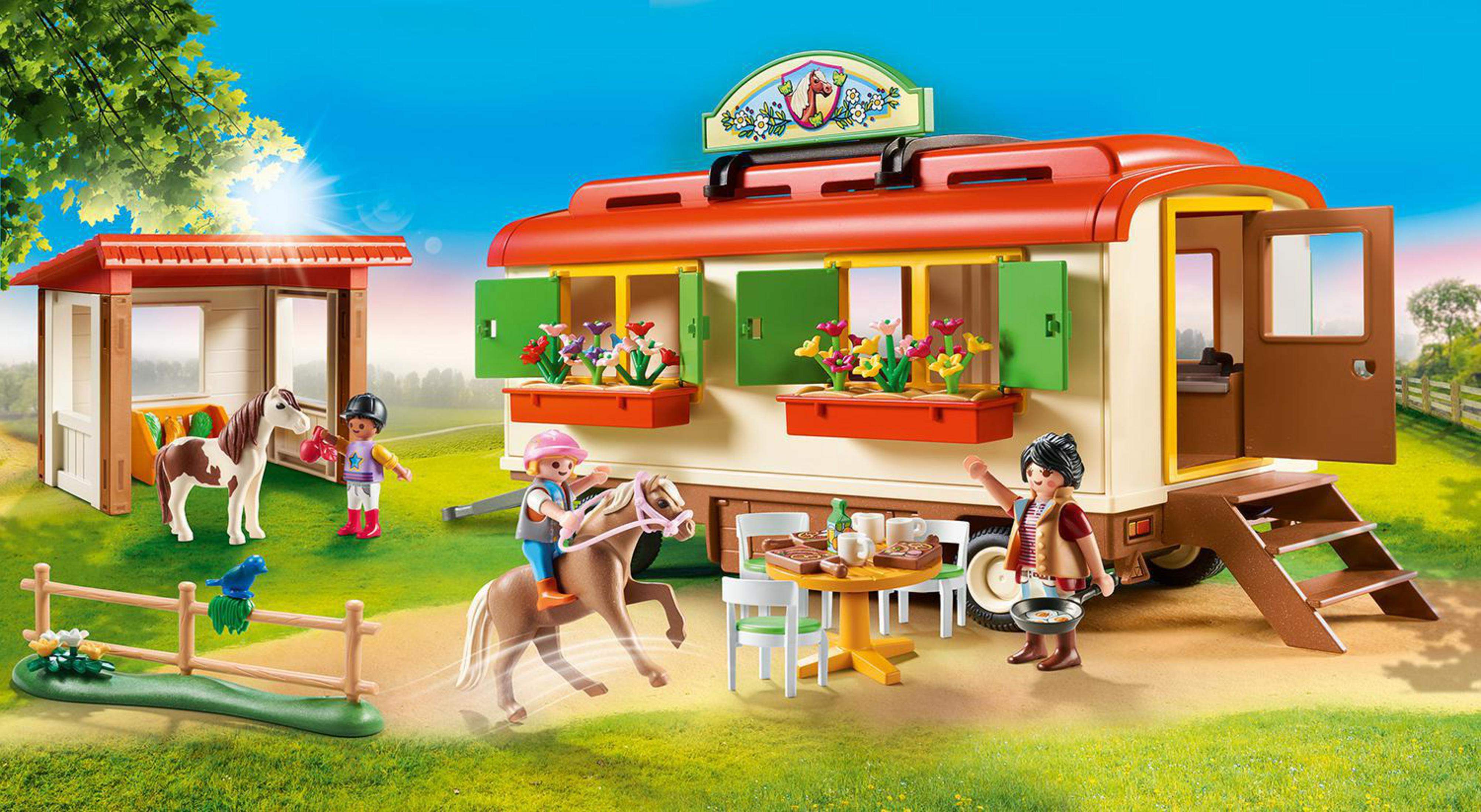 Una scena Playmobil di un rimorchio, cavalli e personaggi. Una ragazza cavalca un cavallo e un'altra cucina a un tavolo.