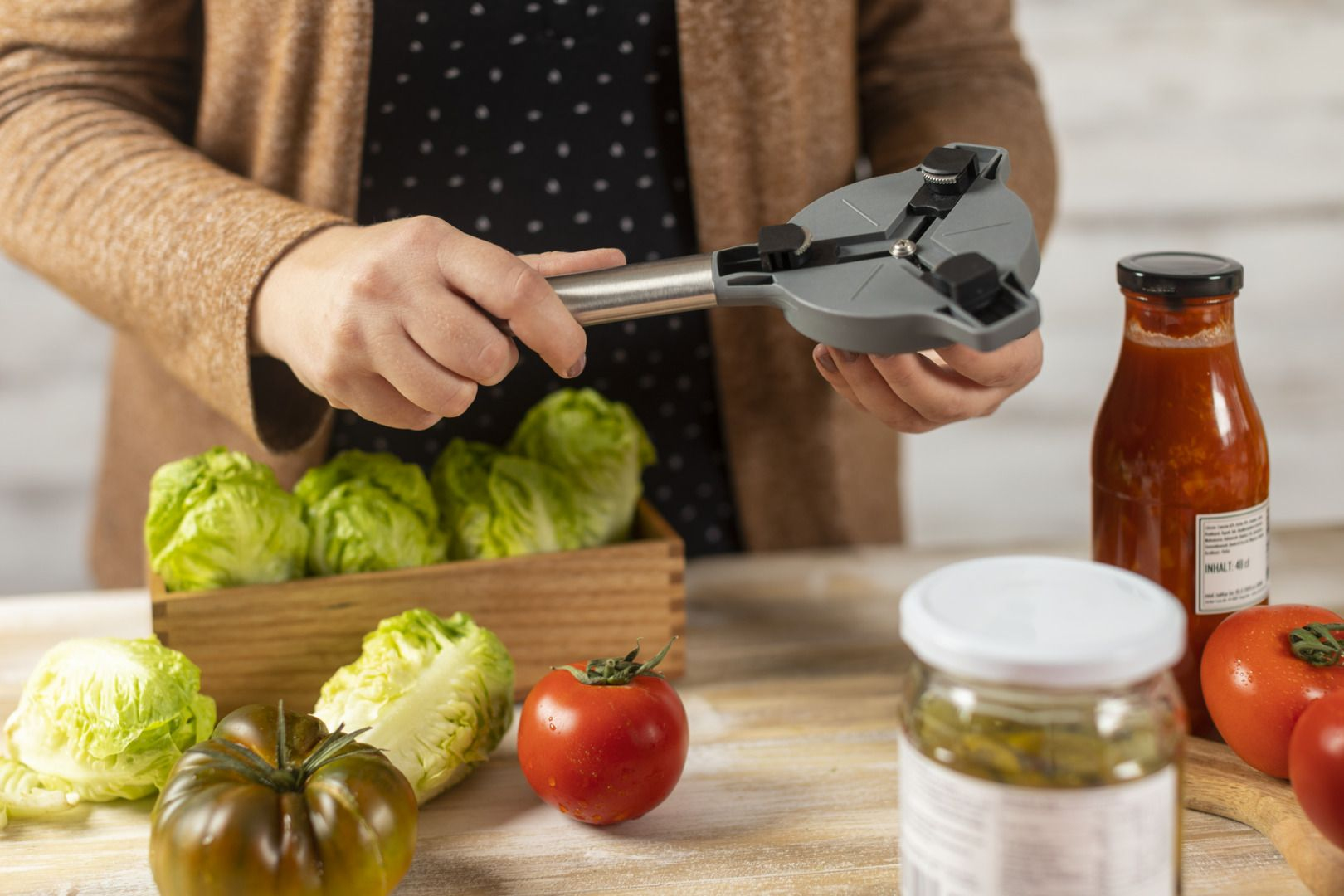 Mani che tengono un apriscatole, con lattuga, pomodori e bottiglie di salsa sullo sfondo.