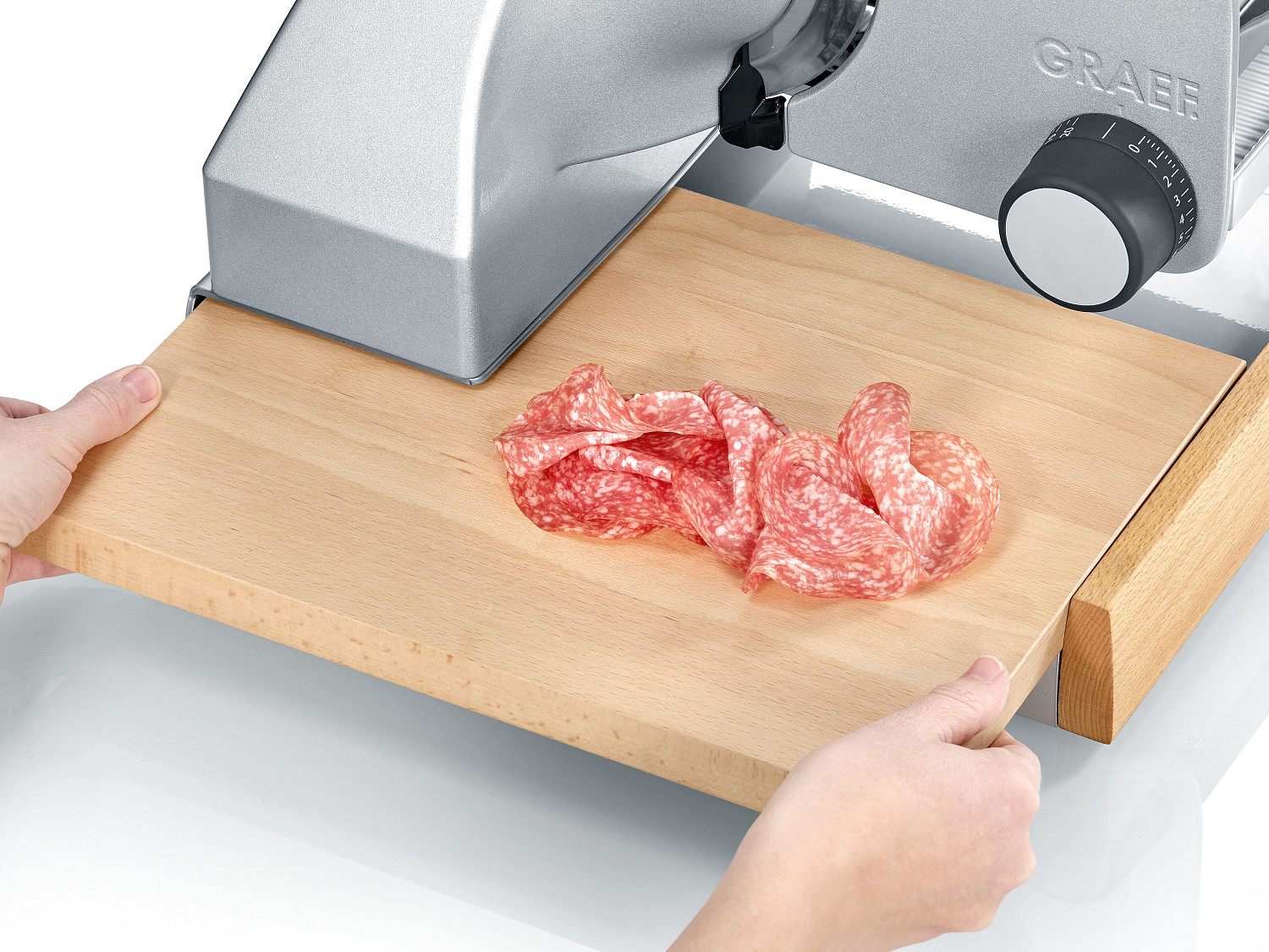 Mains tenant un plateau en bois avec du salami tranché. Trancheuse à viande en arrière-plan.