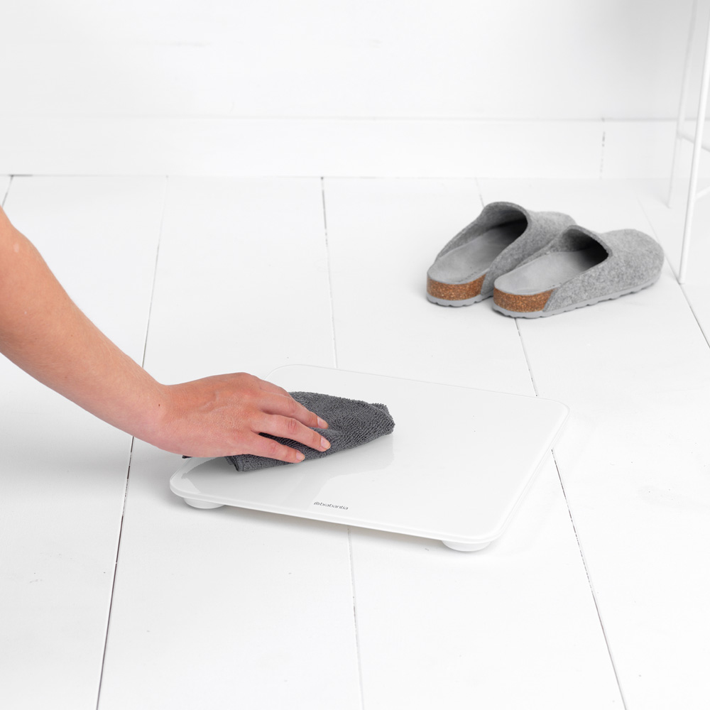 Eine Hand wischt eine weiße Digitalwaage mit einem grauen Tuch ab. Graue Hausschuhe liegen auf dem Boden.