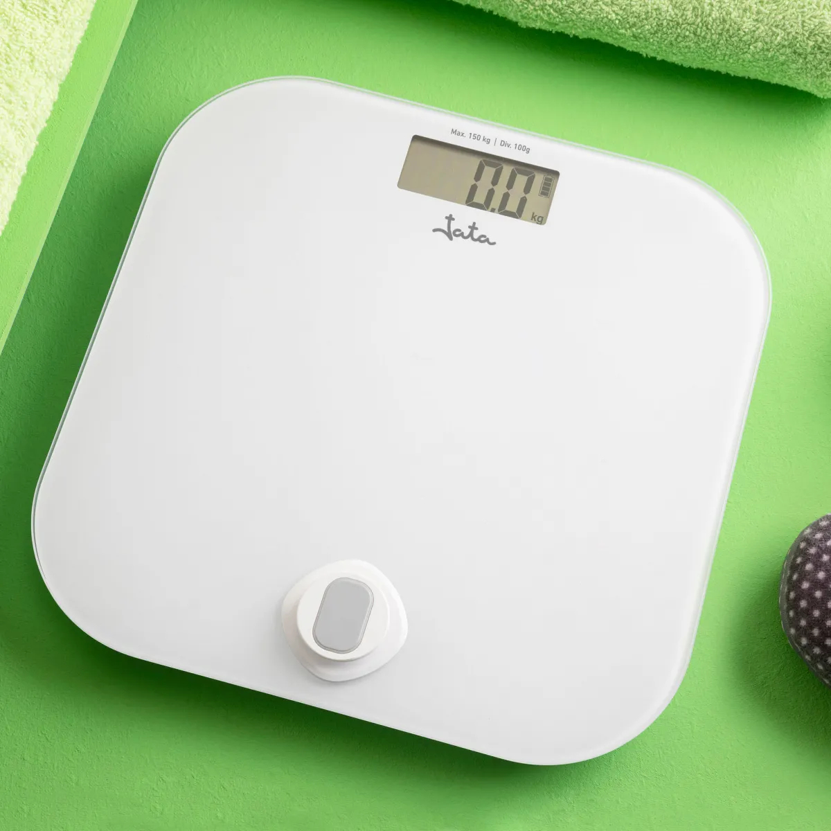 Biała waga cyfrowa pokazuje 0,0 kg na zielonej powierzchni, obok zielony ręcznik.