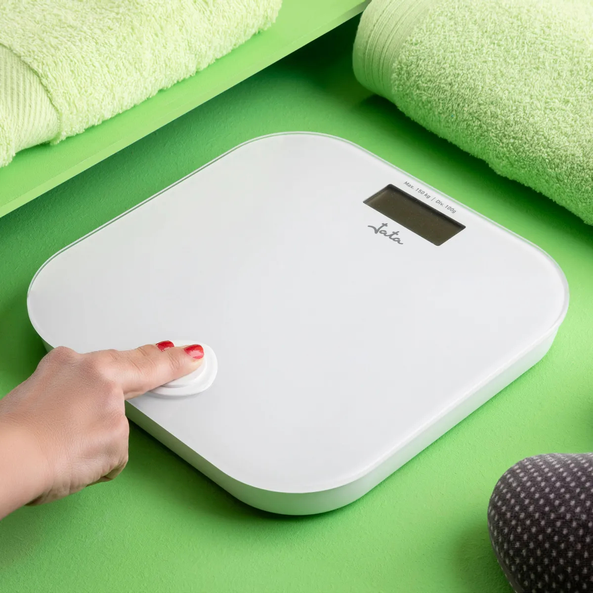 Biała waga cyfrowa na zielonej powierzchni, dłoń naciska przycisk. Widoczny zielony ręcznik.