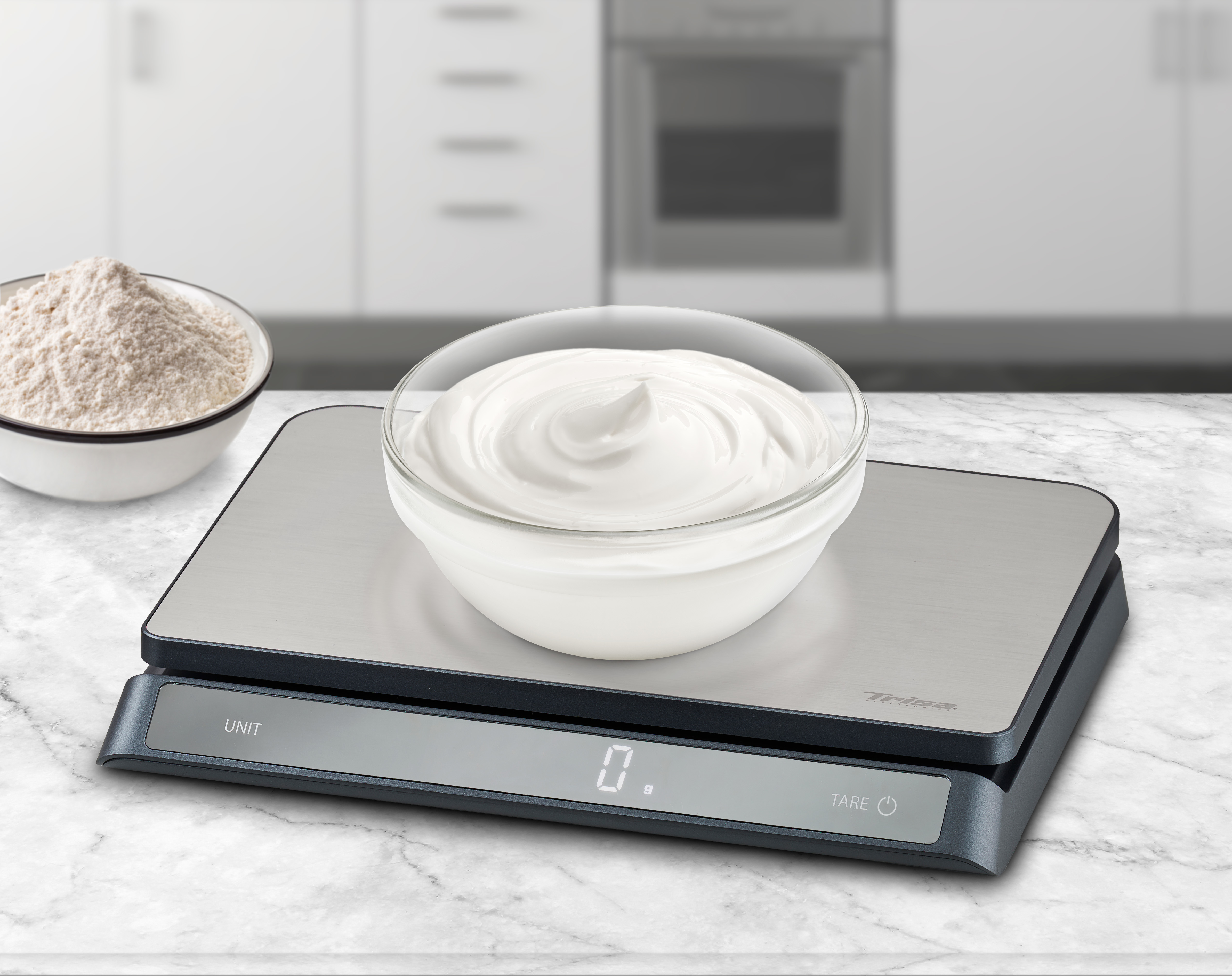 Küchenwaage mit einer Schüssel weißer Creme und einer Schüssel Mehl. Neutraler Küchenhintergrund.