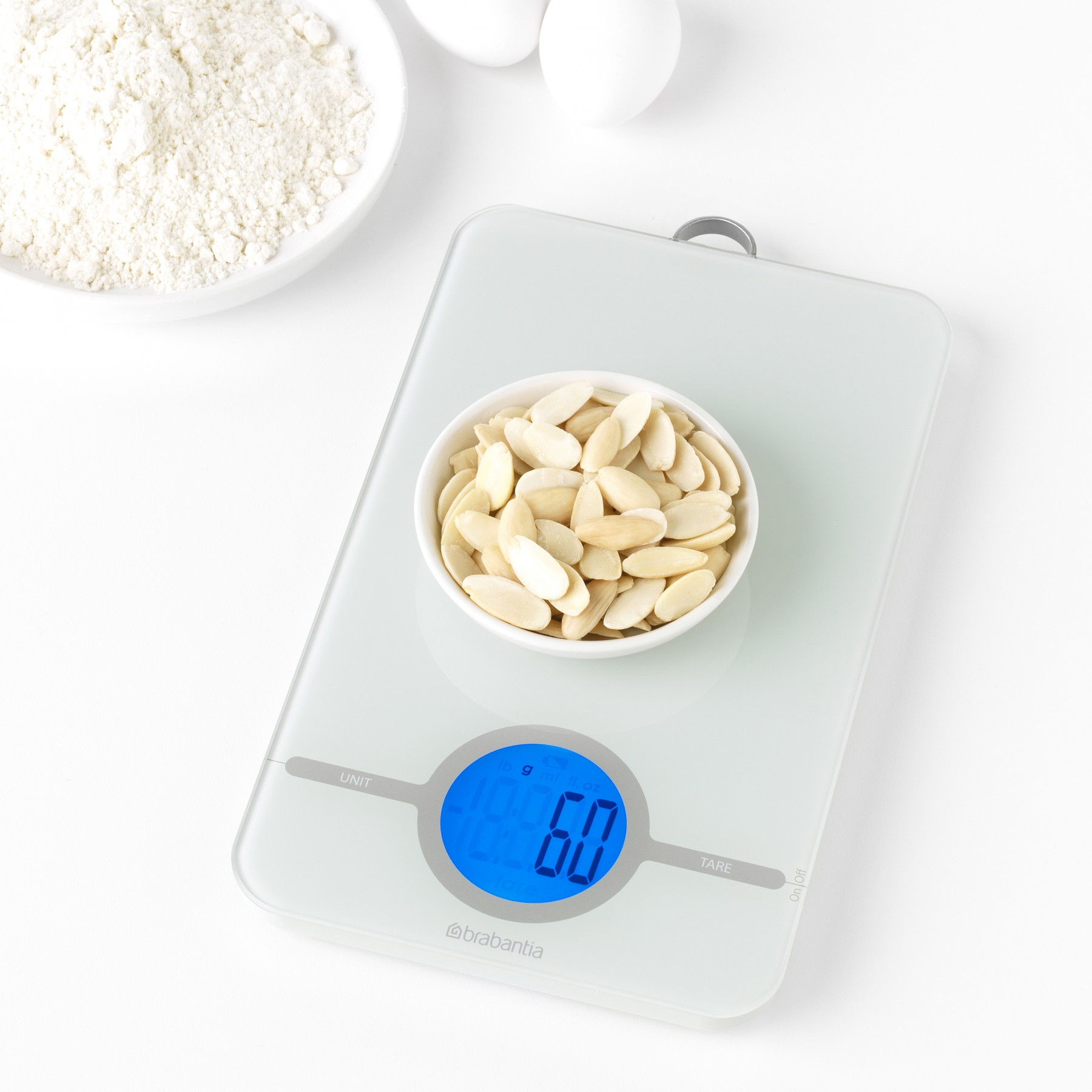 Biała waga cyfrowa z miską migdałów, mąką i dwoma jajkami na białej powierzchni.