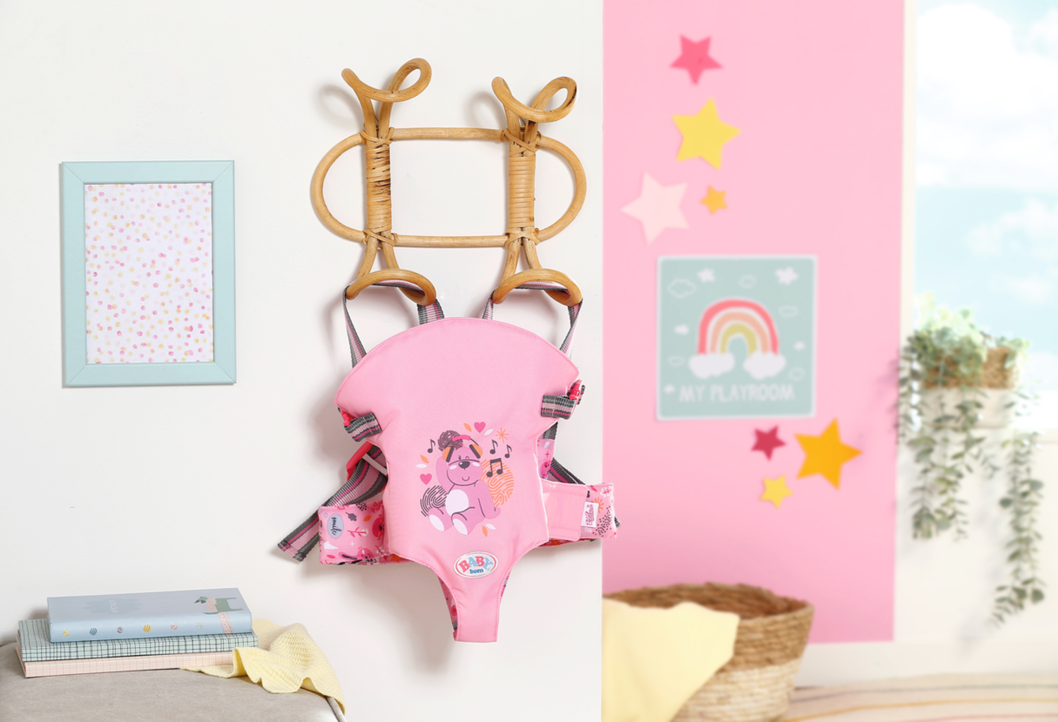 Różowy nosidełko dla dziecka wiszące na drewnianym wieszaku. Ramka na zdjęcia i gwiazdy zdobią różową ścianę.