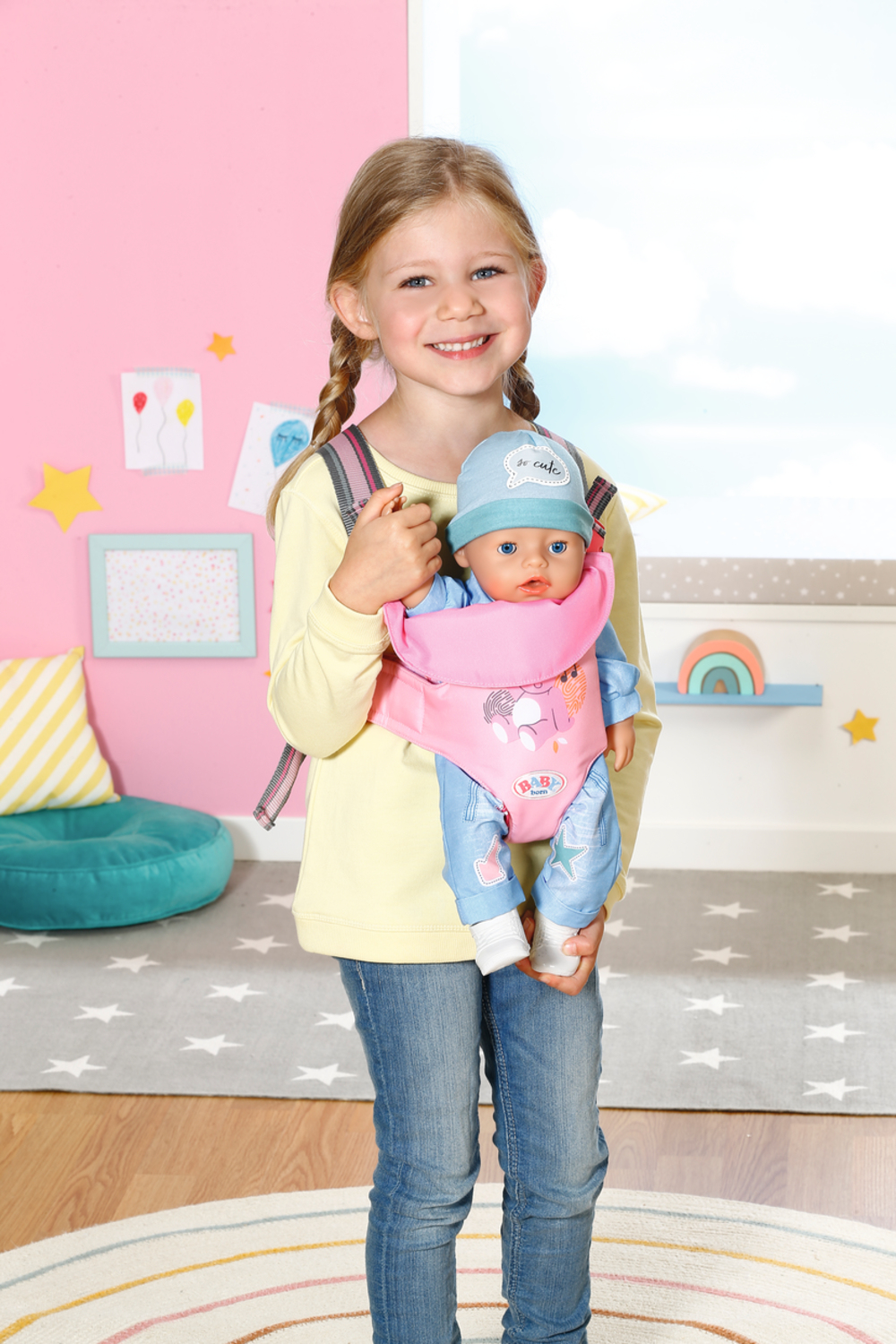 Dziewczyna z warkoczami, żółtej koszulce i dżinsach, trzymająca lalkę w nosidełku.