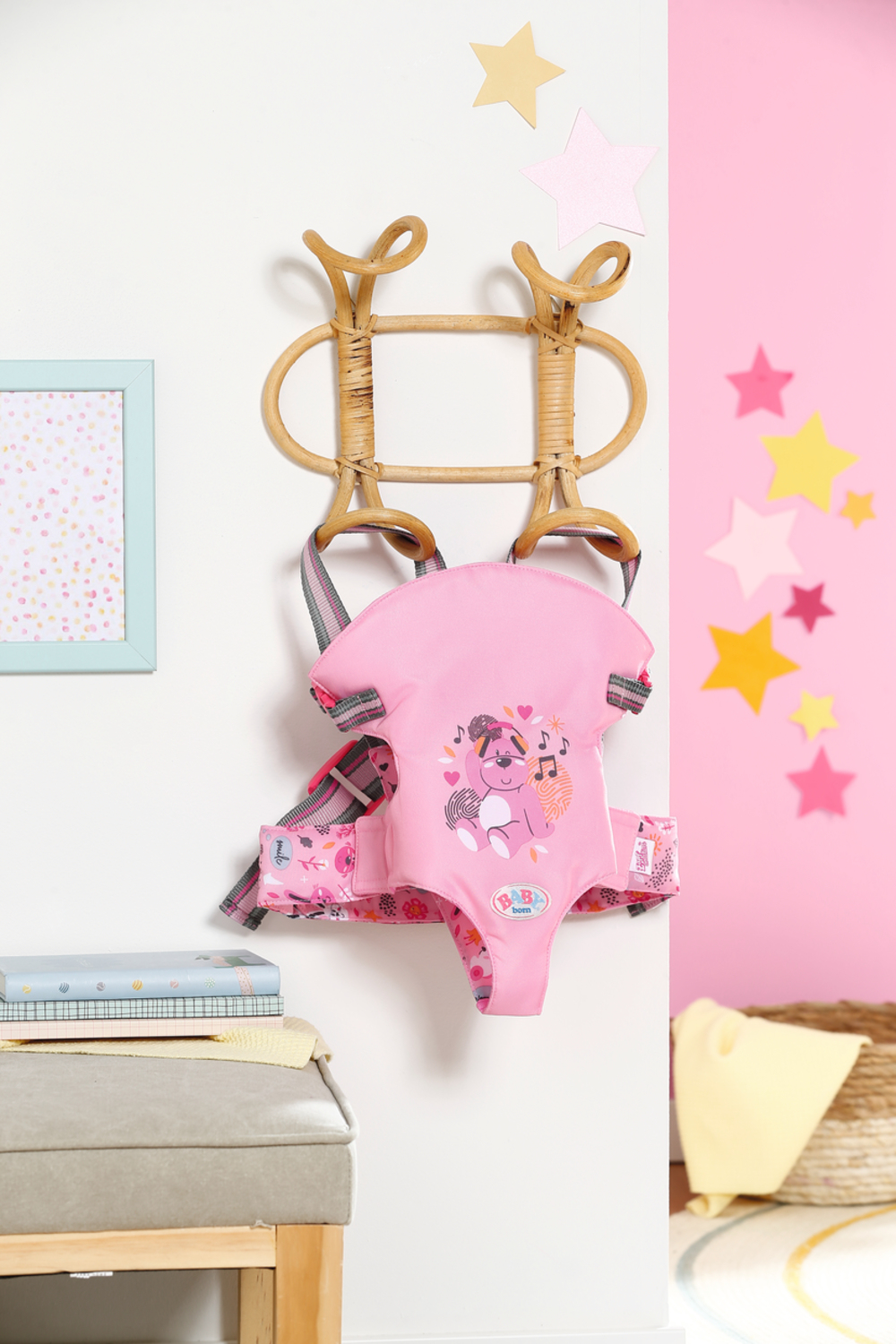 Różowy nosidełko dla dziecka wiszące na ścianie. Ma wzór z kreskówek i obok jest oprawiony obrazek.