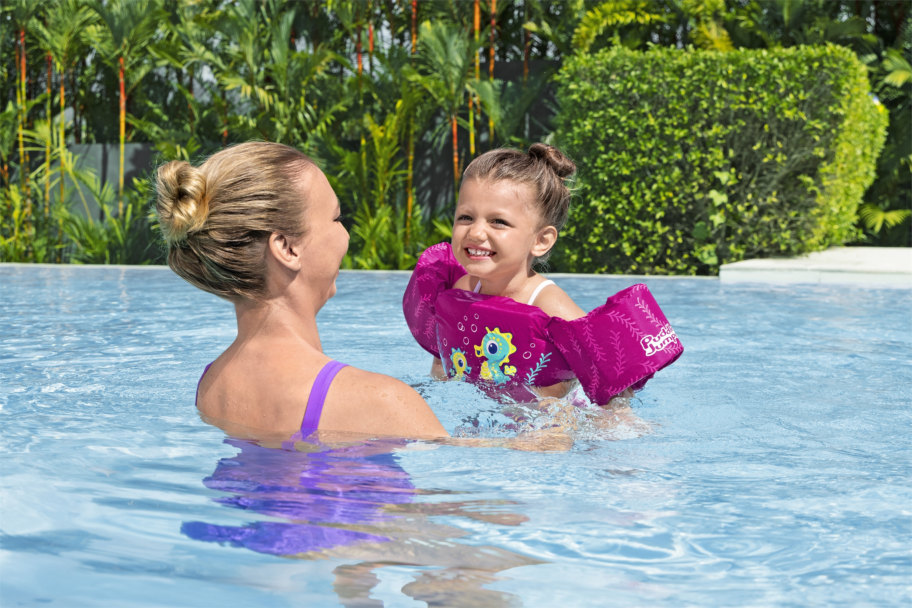 Donna in piscina con bambina con braccioli rosa, sorridente. Cespugli verdi sullo sfondo, acqua blu.