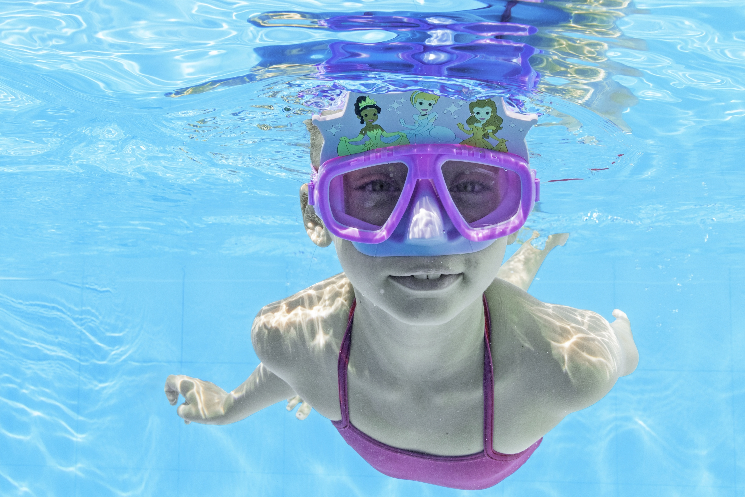 Una ragazza con un costume da bagno rosa e una maschera a tema principessa nuota sott'acqua, guardando verso la fotocamera.