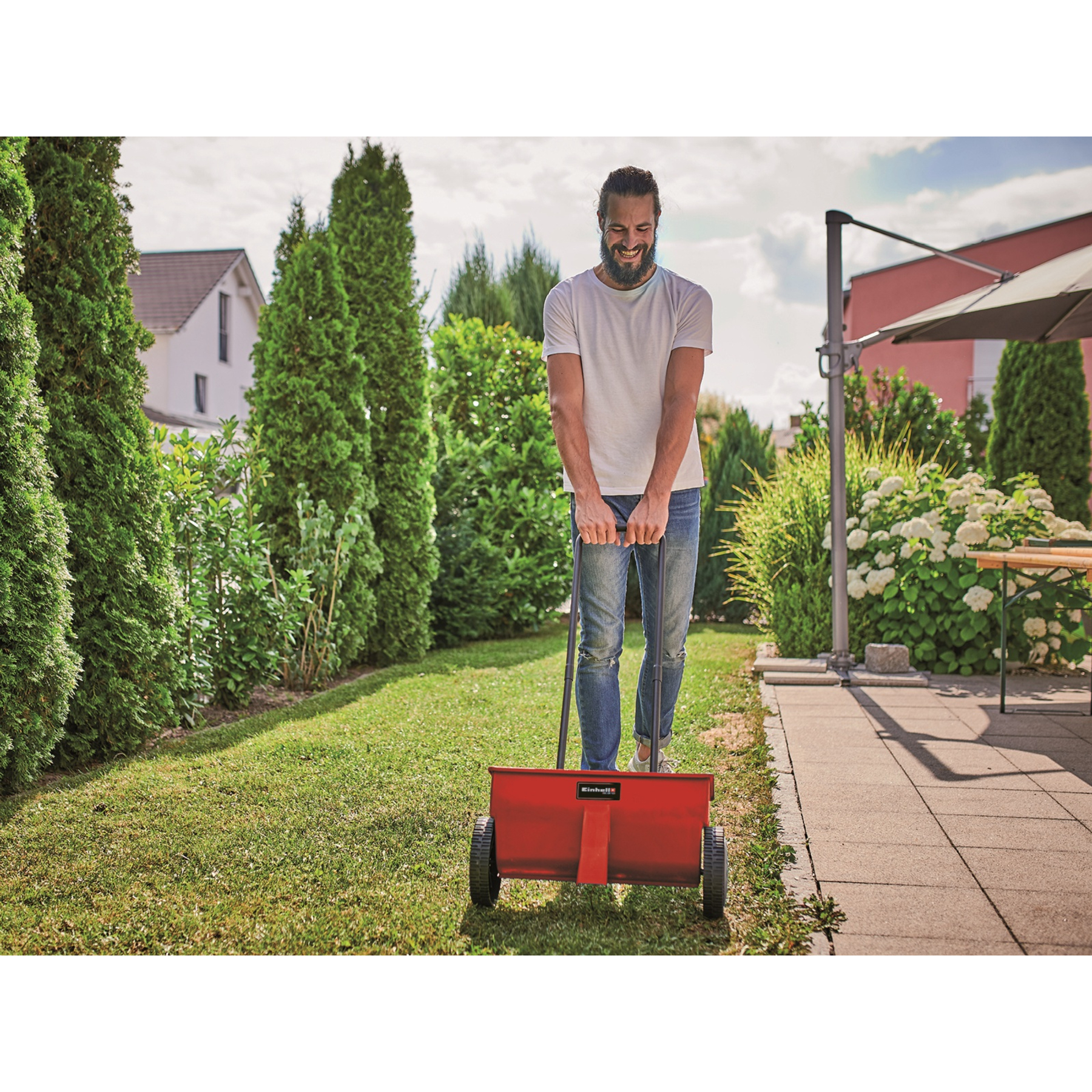 Mężczyzna pcha czerwony rozsiewacz na zielonym trawniku, otoczonym krzewami i drzewami.
