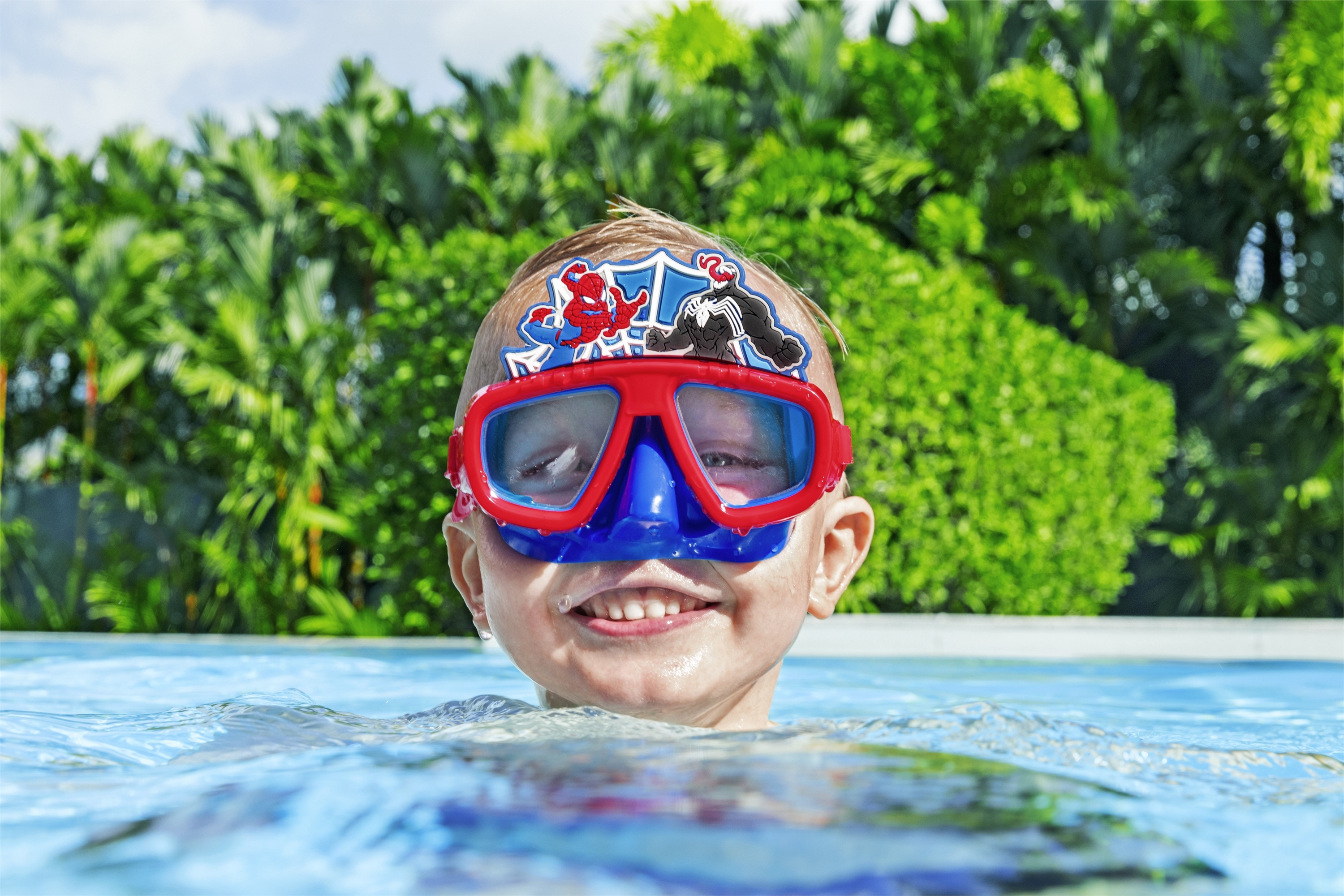 Ragazzo in acqua con occhialini di Spider-Man, sorridente, con vegetazione verde.