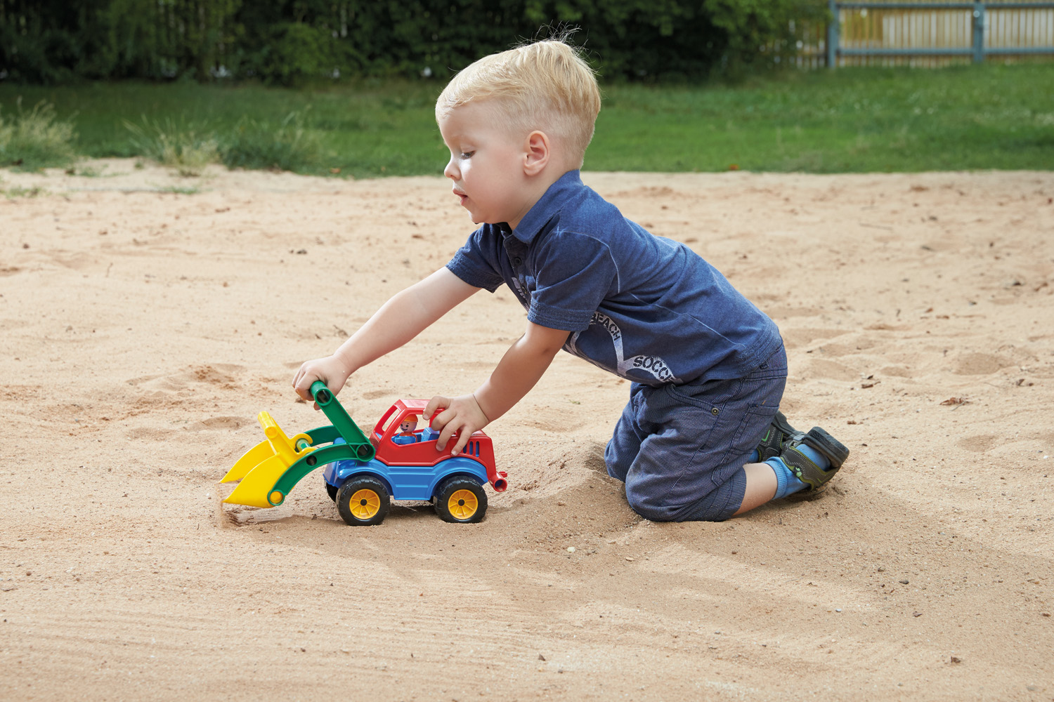 Młody, blond włosy chłopiec bawiący się w piasku z czerwoną, niebieską i żółtą koparką.