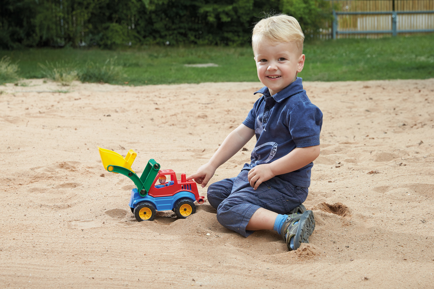 Chłopiec siedzi w piasku, bawiąc się czerwoną, niebieską i żółtą ciężarówką zabawkową.