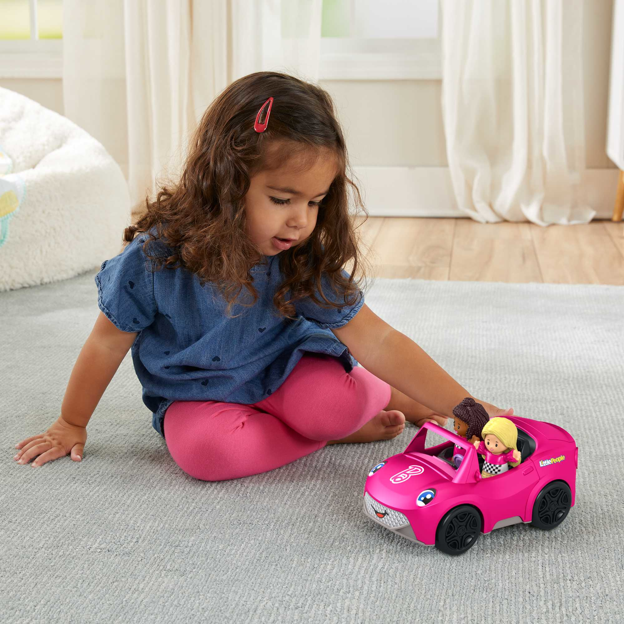 Una niña juega con un coche de juguete rosa con dos muñecas dentro. La escena es en interiores.