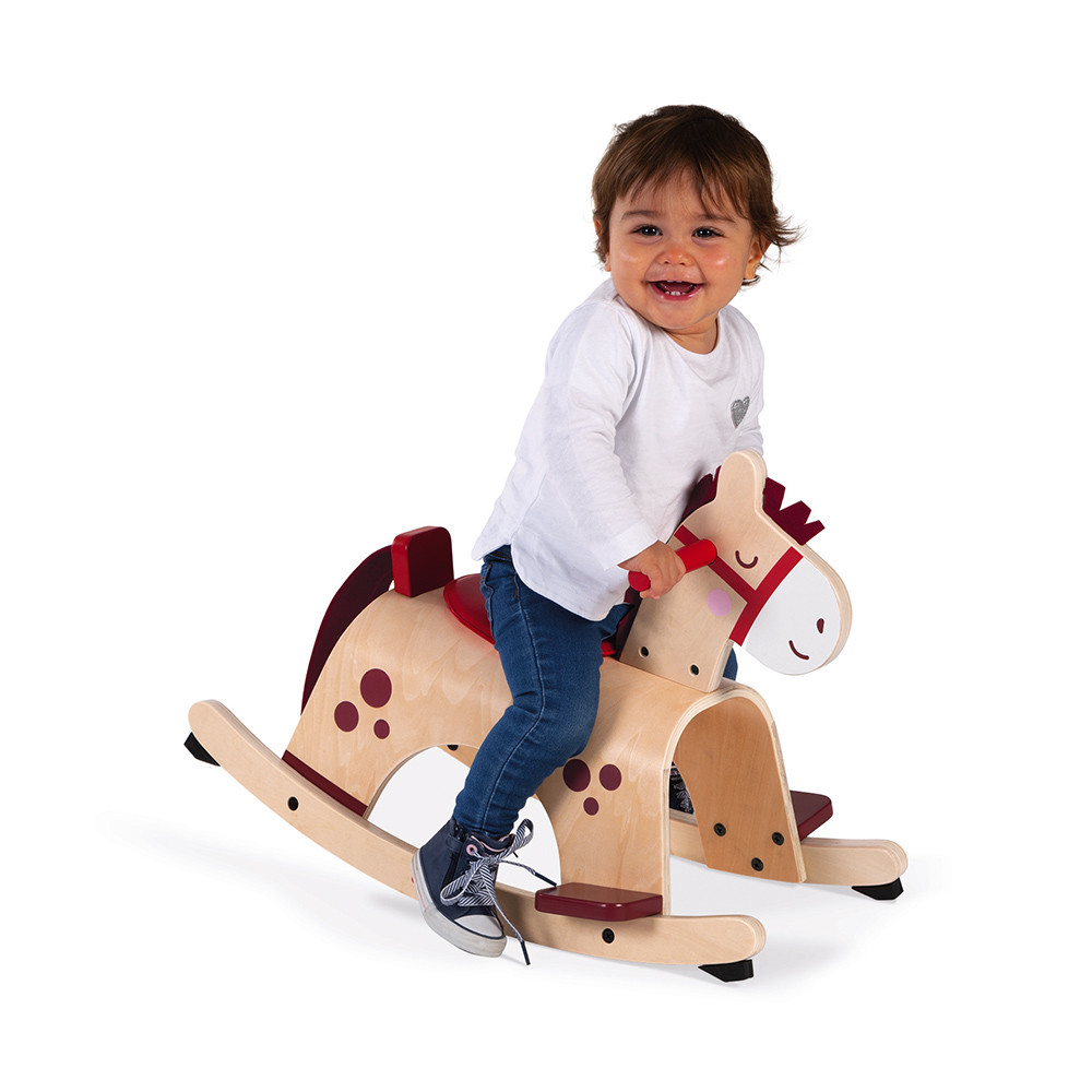Ein Baby auf einem hölzernen Schaukelpferd, das lächelt, Jeans und ein weißes Hemd trägt, auf Weiß.