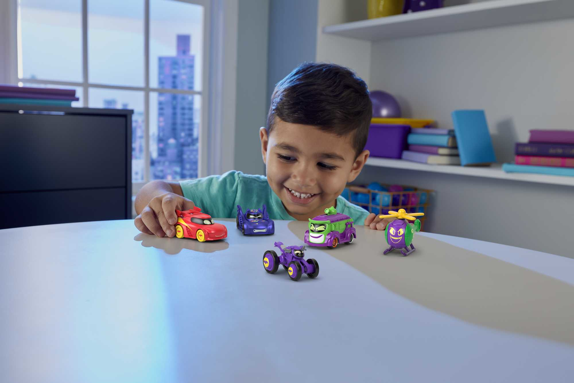 Chłopiec uśmiecha się do pięciu zabawek-samochodów na stole. Samochody mają różne kolory i wzory.