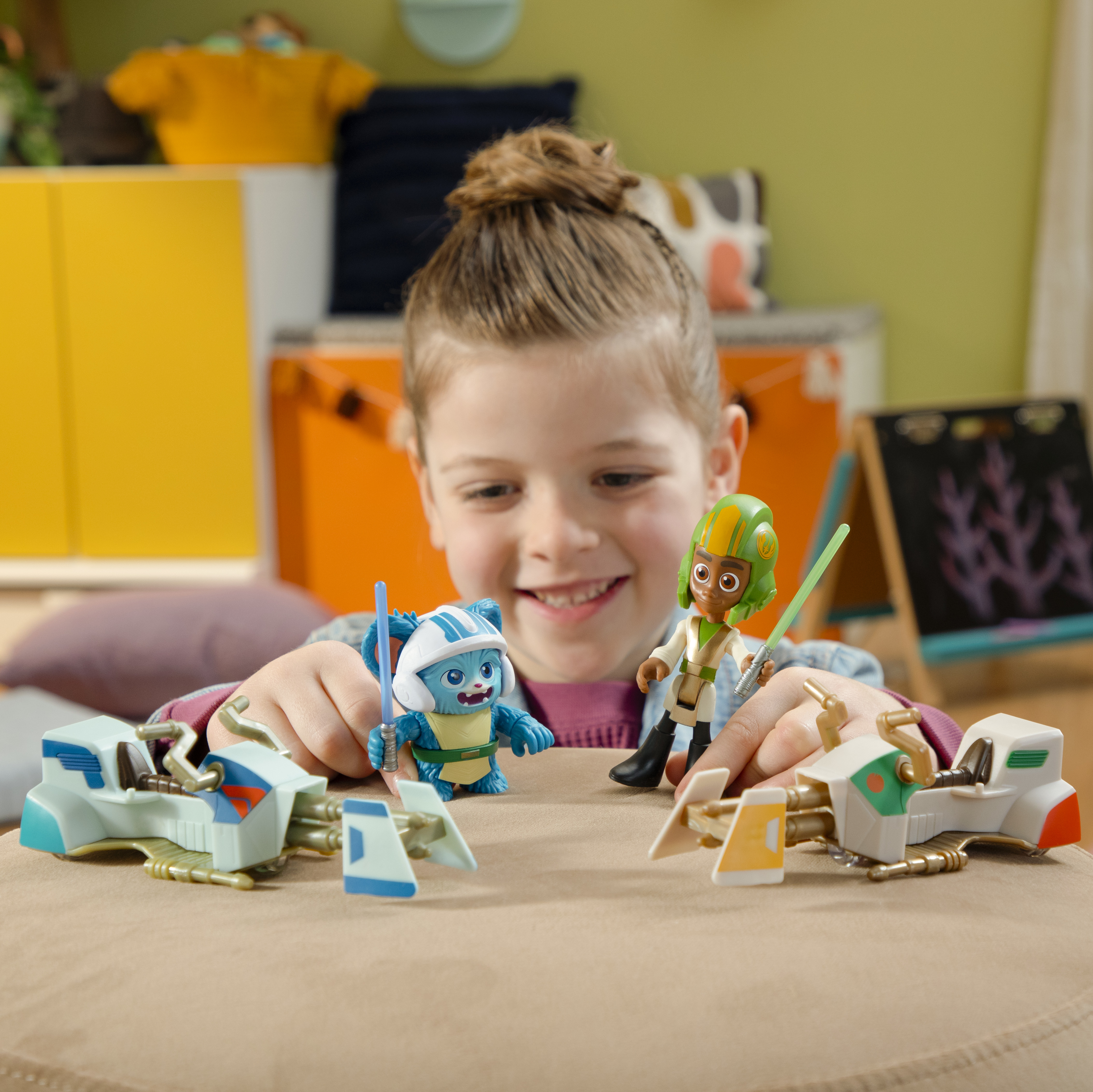 Mała dziewczynka bawi się zabawkami z Gwiezdnych Wojen. Dwie postacie z mieczami świetlnymi i dwa ścigacze na stole.