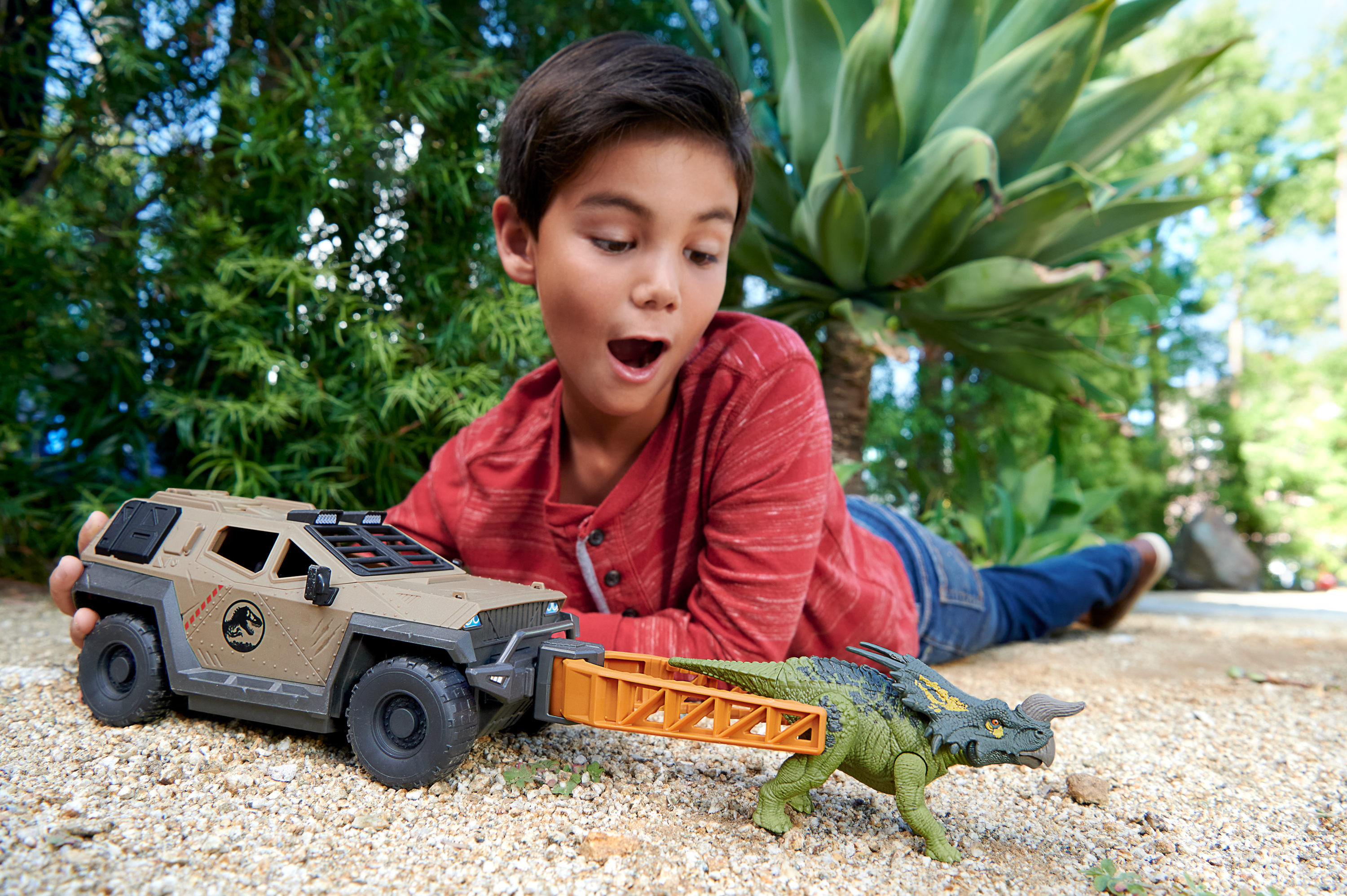 Ein Junge spielt mit einem Spielzeug-SUV und einem Dinosaurier-Spielzeug. Die Szene ist im Freien mit einem verschwommenen Hintergrund aus grünen Pflanzen.