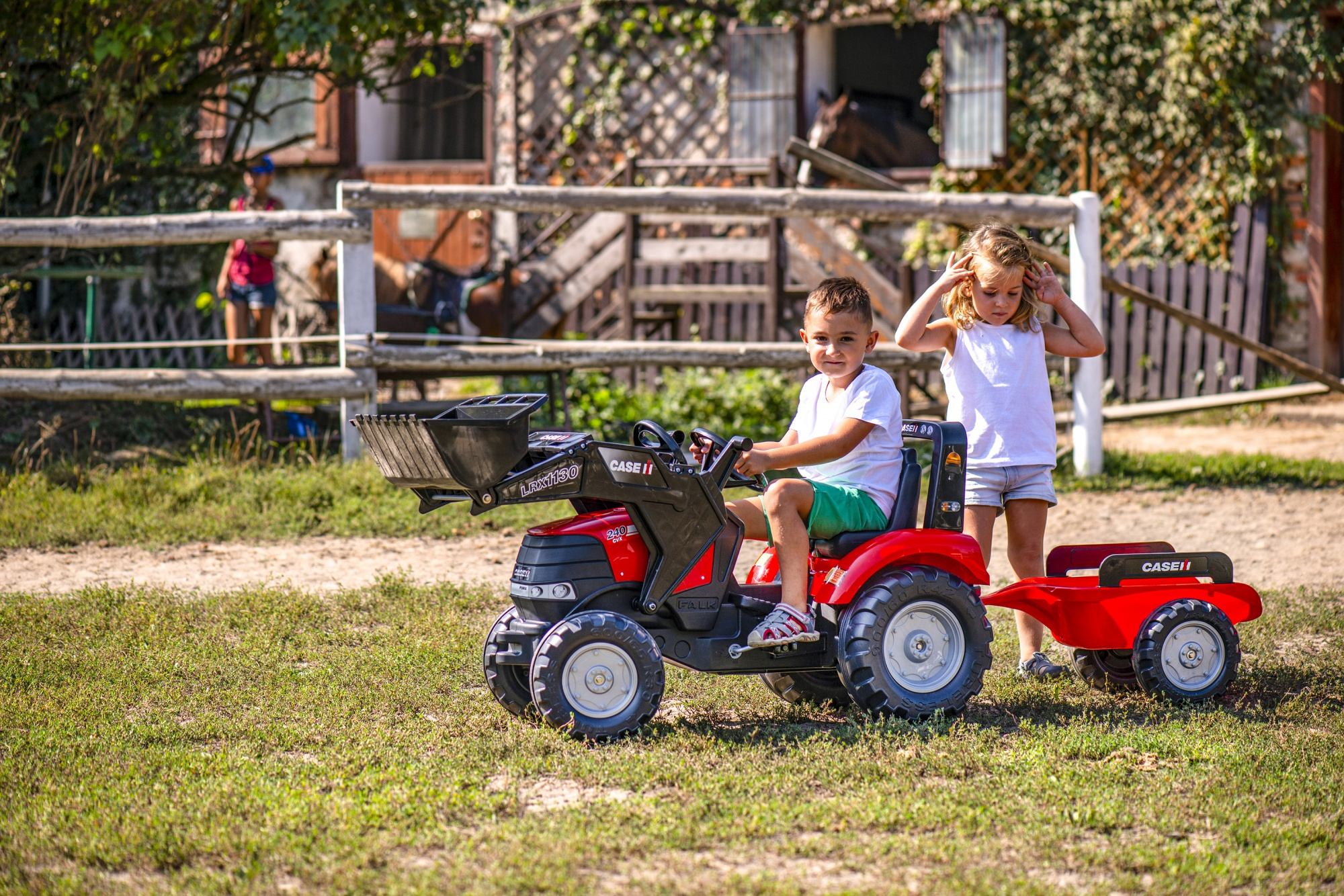 Dos niños jugando con un tractor rojo con remolque en un campo verde.