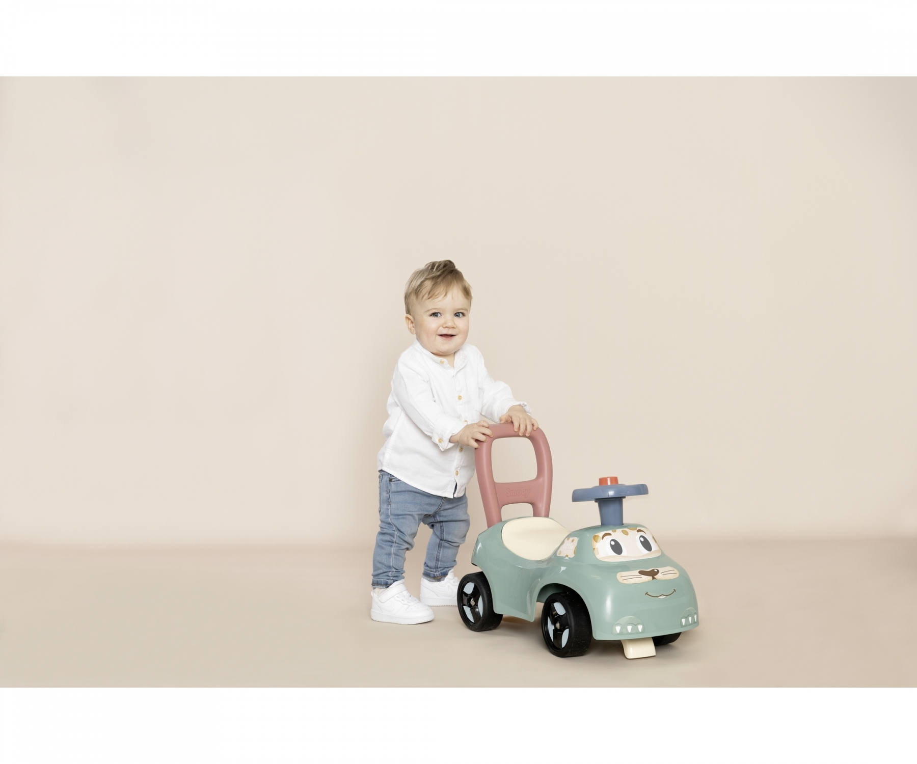 Un bambino piccolo sta spingendo un'auto giocattolo verde e rosa. Sono in piedi su un pavimento beige.