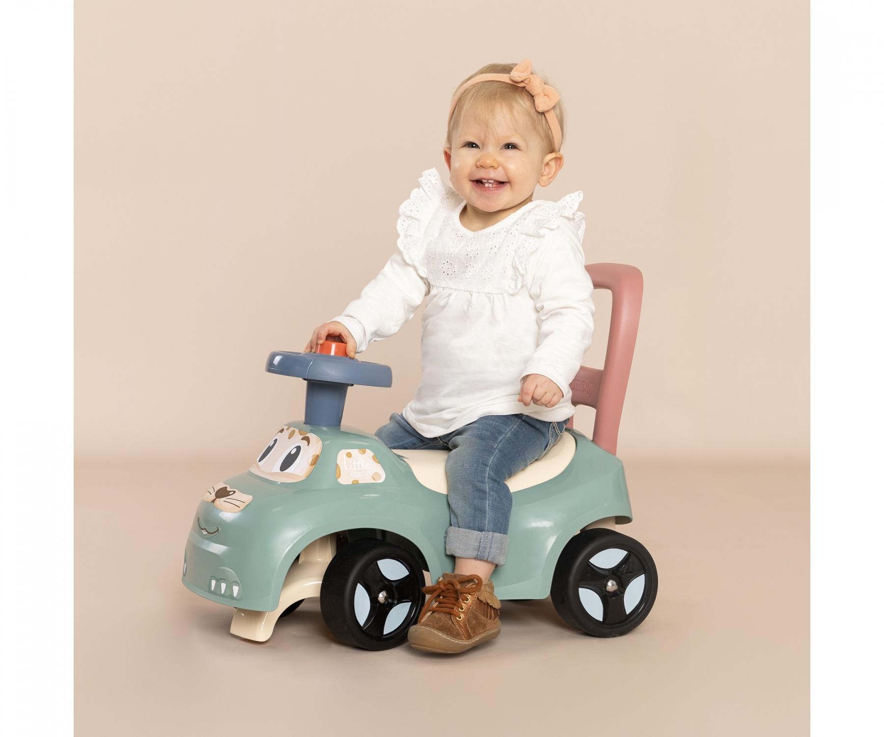 Bambina seduta su un'auto giocattolo blu e bianca che sorride, sfondo pesca, scarpe marroni.
