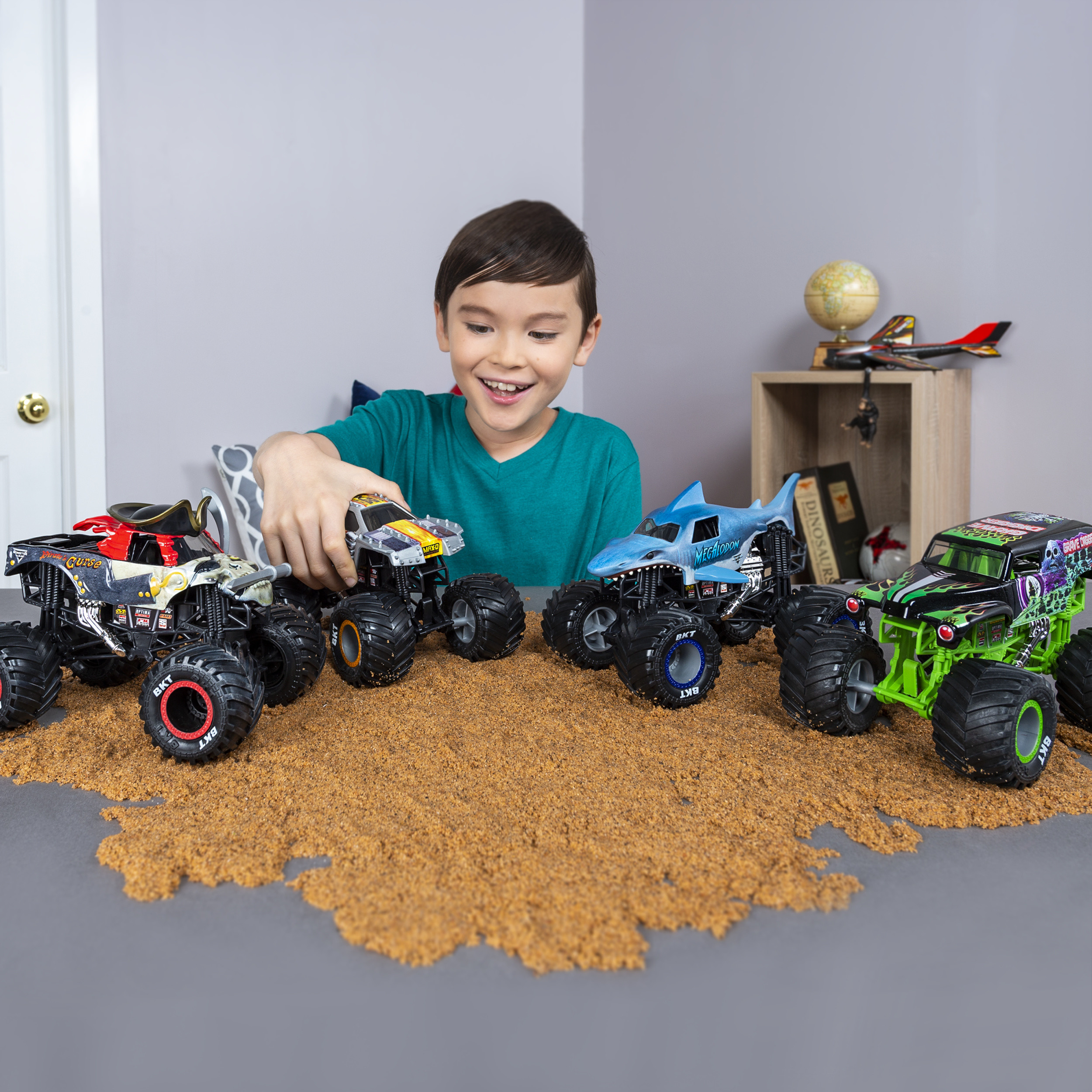 Un niño pequeño jugando con varios camiones monstruo en una superficie arenosa.
