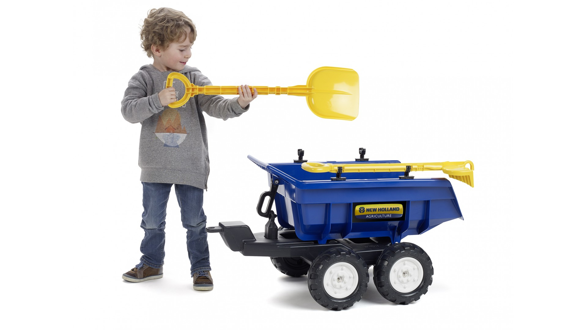 Ein kleiner Junge mit einer gelben Schaufel neben einem blauen Spielzeugtraktor mit Anhänger.