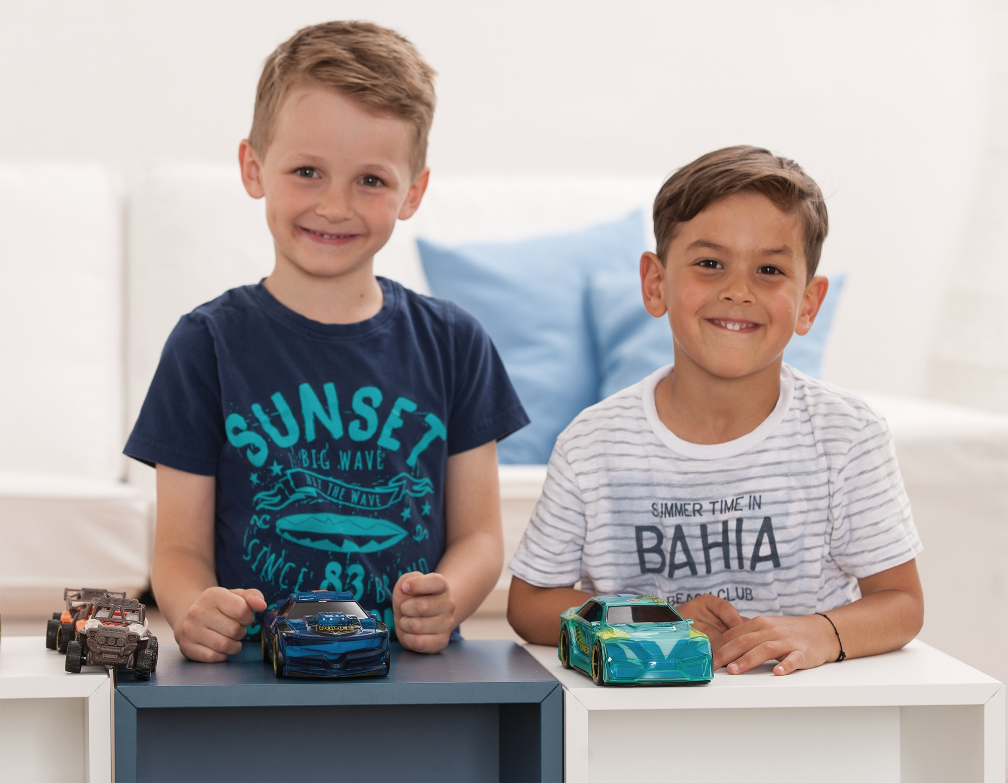 Zwei Jungen stehen zusammen und lächeln, mit Spielzeugautos auf einem Tisch vor ihnen. Heller Hintergrund.