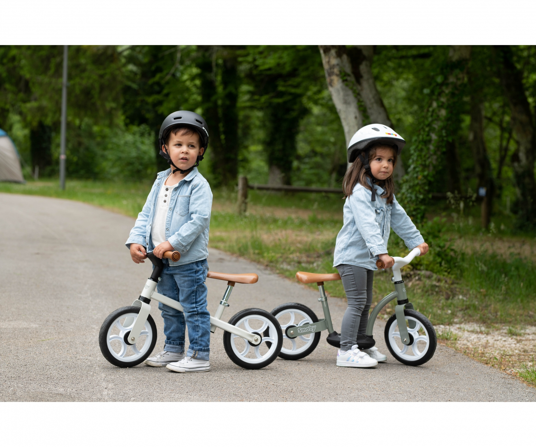 Due bambini su bici senza pedali, con caschi. Sono in un parco con uno sfondo verde.