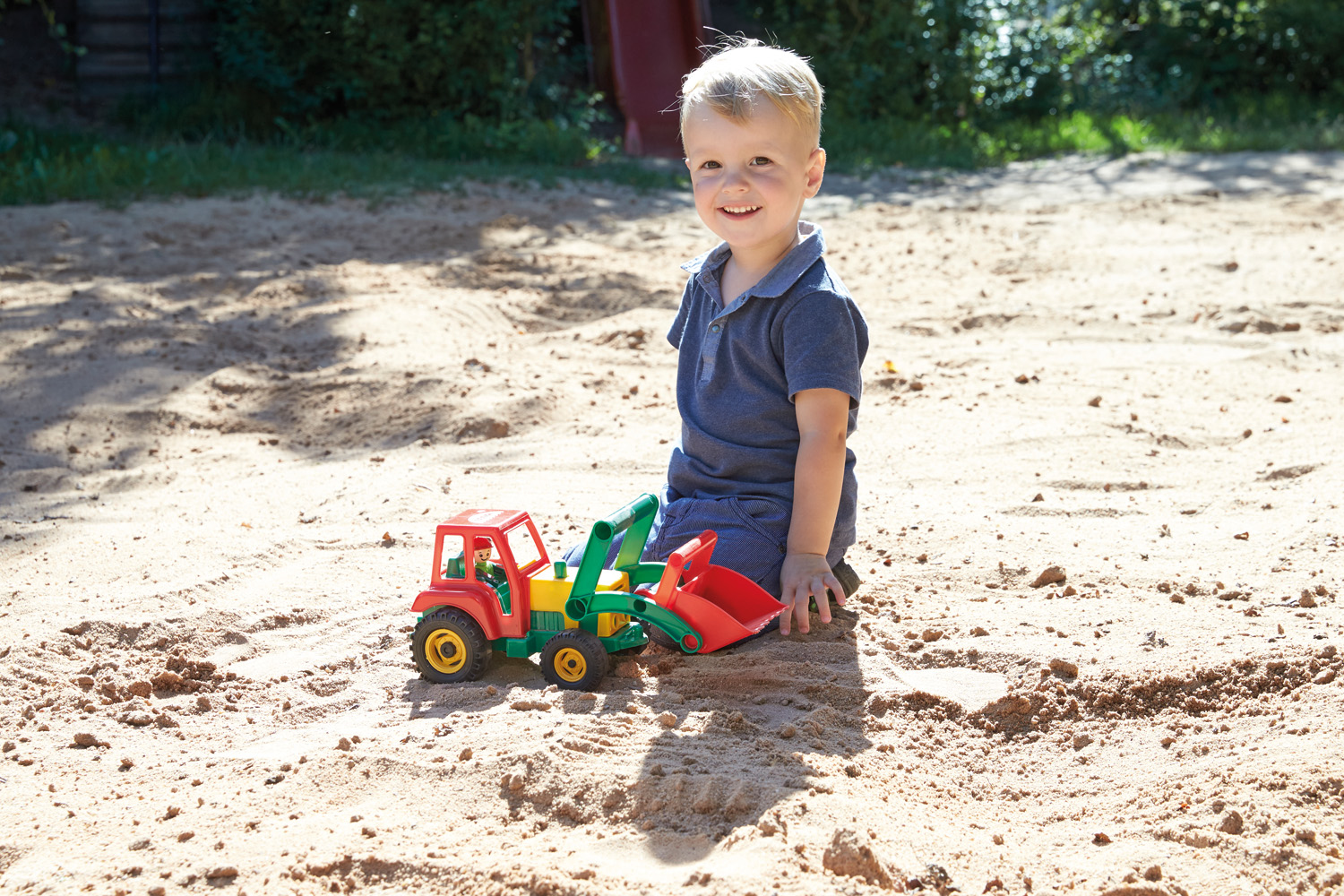 Chłopiec siedzi w piaskownicy, bawiąc się czerwoną, zieloną i żółtą zabawką ciężarówką.