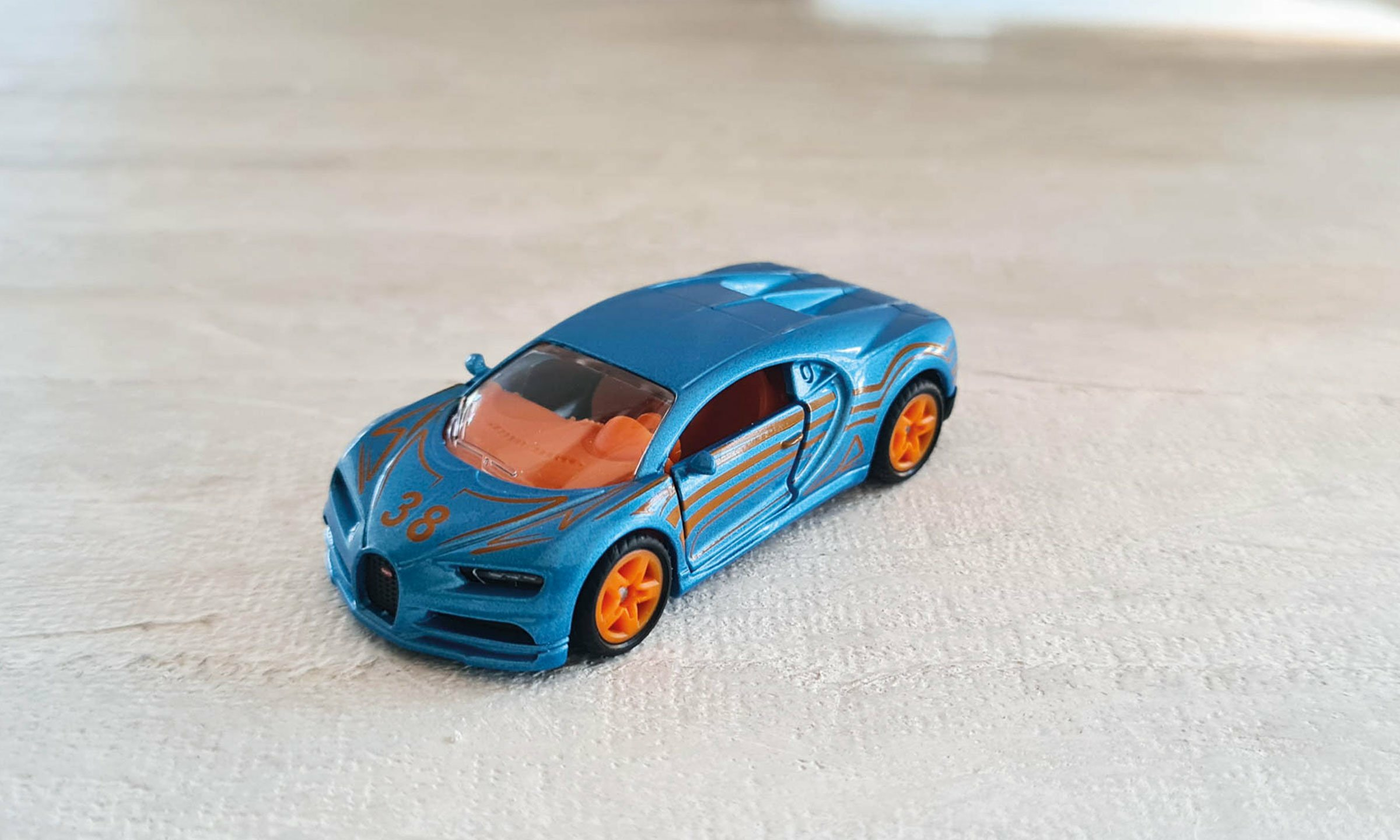 Niebieski samochodzik z pomarańczowymi akcentami. Samochód stoi na białej drewnianej powierzchni.