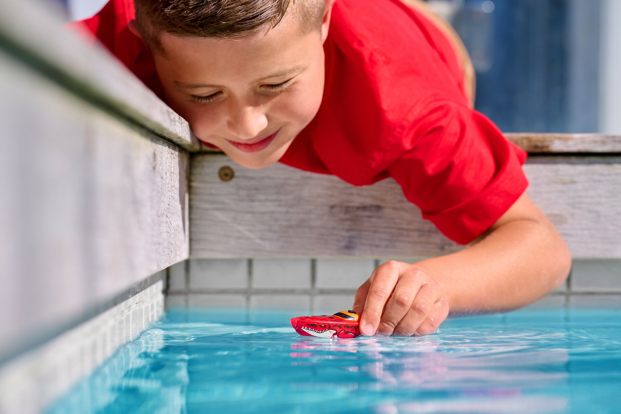 Chłopiec sięgający do wody, aby bawić się małą czerwoną zabawką łodzią w basenie.