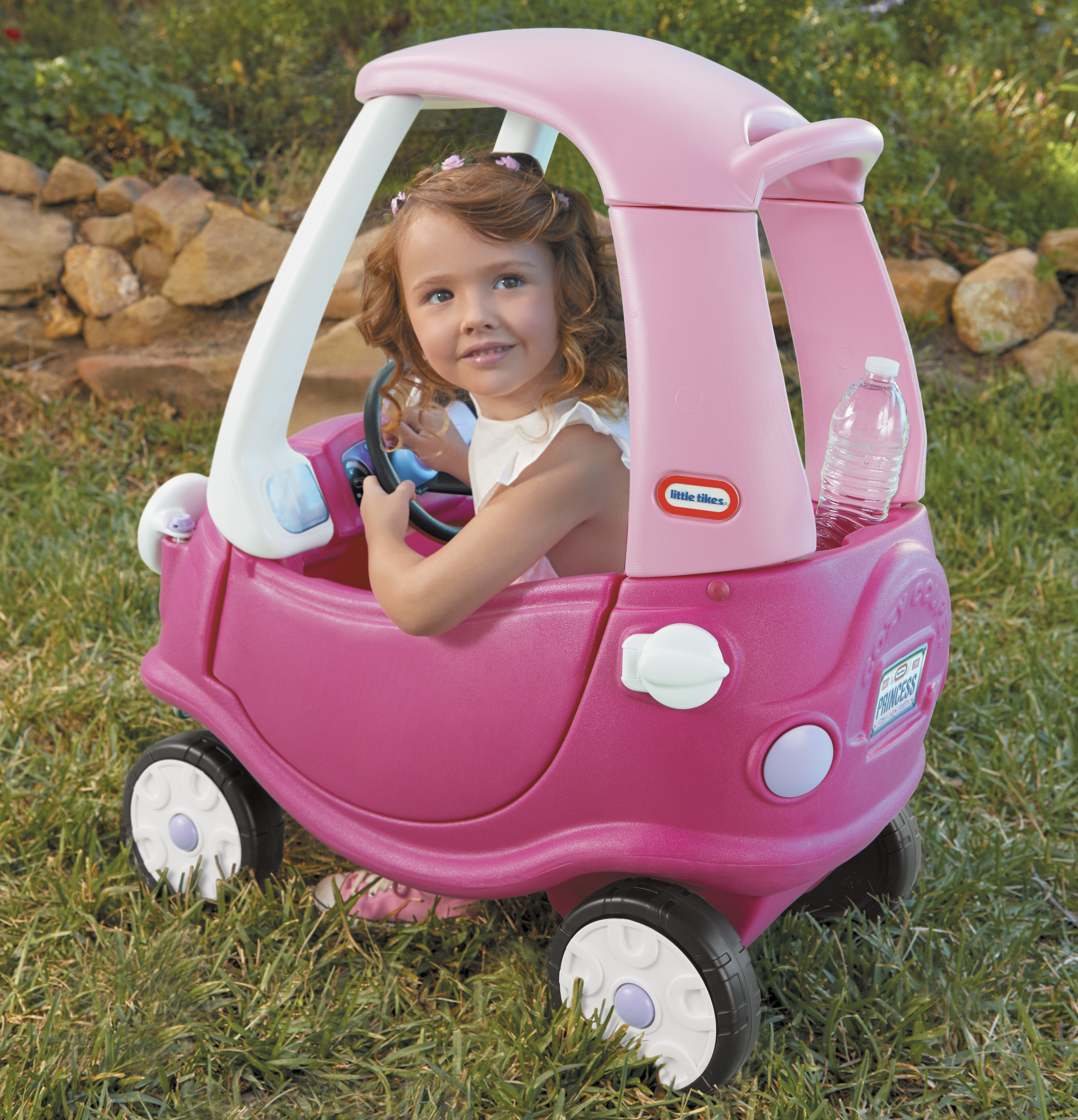 Una bambina è in un'auto giocattolo rosa, sorridendo mentre è seduta sull'erba con una bottiglia d'acqua.
