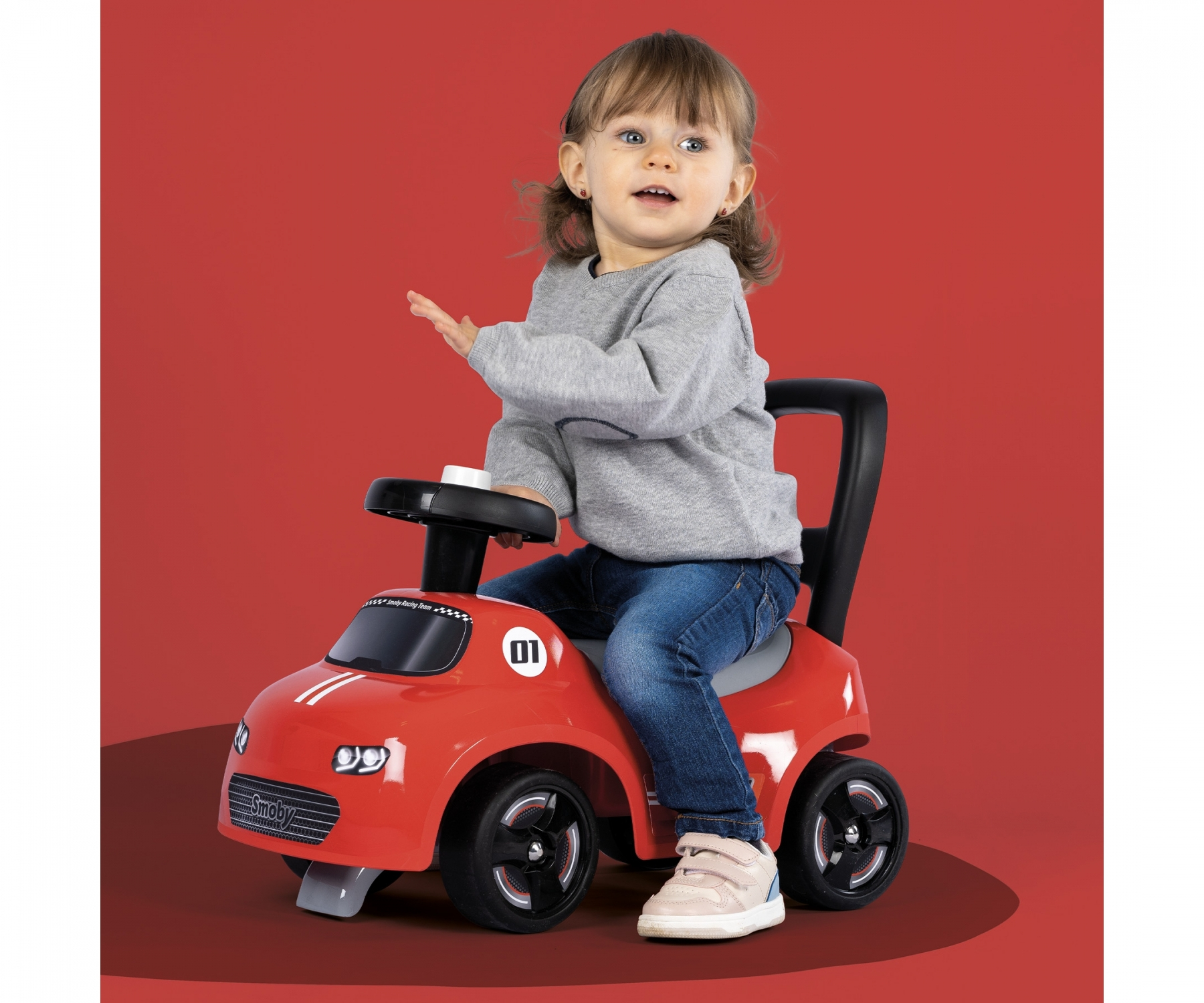 Un bambino piccolo è seduto su un'auto rossa, sorridente, su uno sfondo rosso.