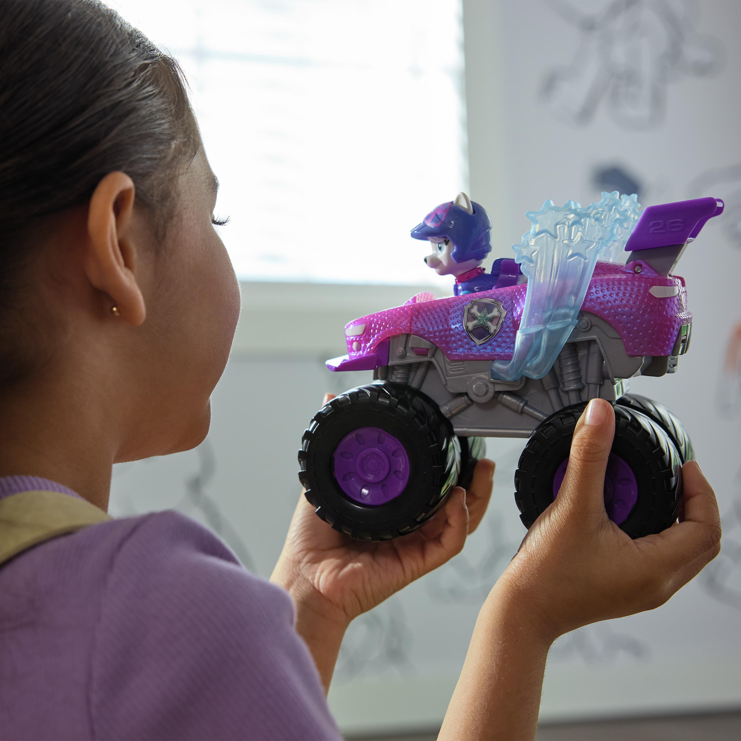 Dziewczyna trzyma fioletową zabawkę w postaci monster trucka. Zabawka ma postać z Psiego Patrolu.
