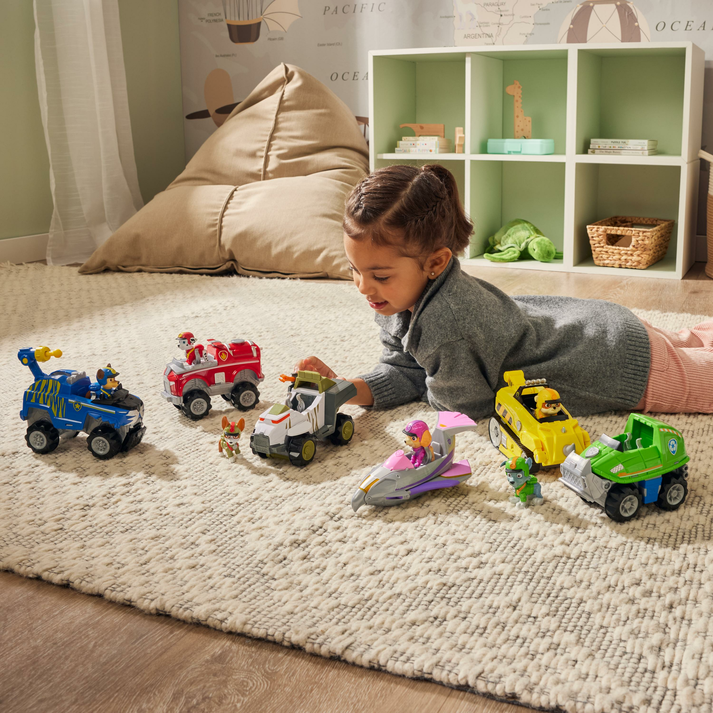 Mała dziewczynka leży na dywanie, bawiąc się pojazdami i figurkami zabawek Paw Patrol.