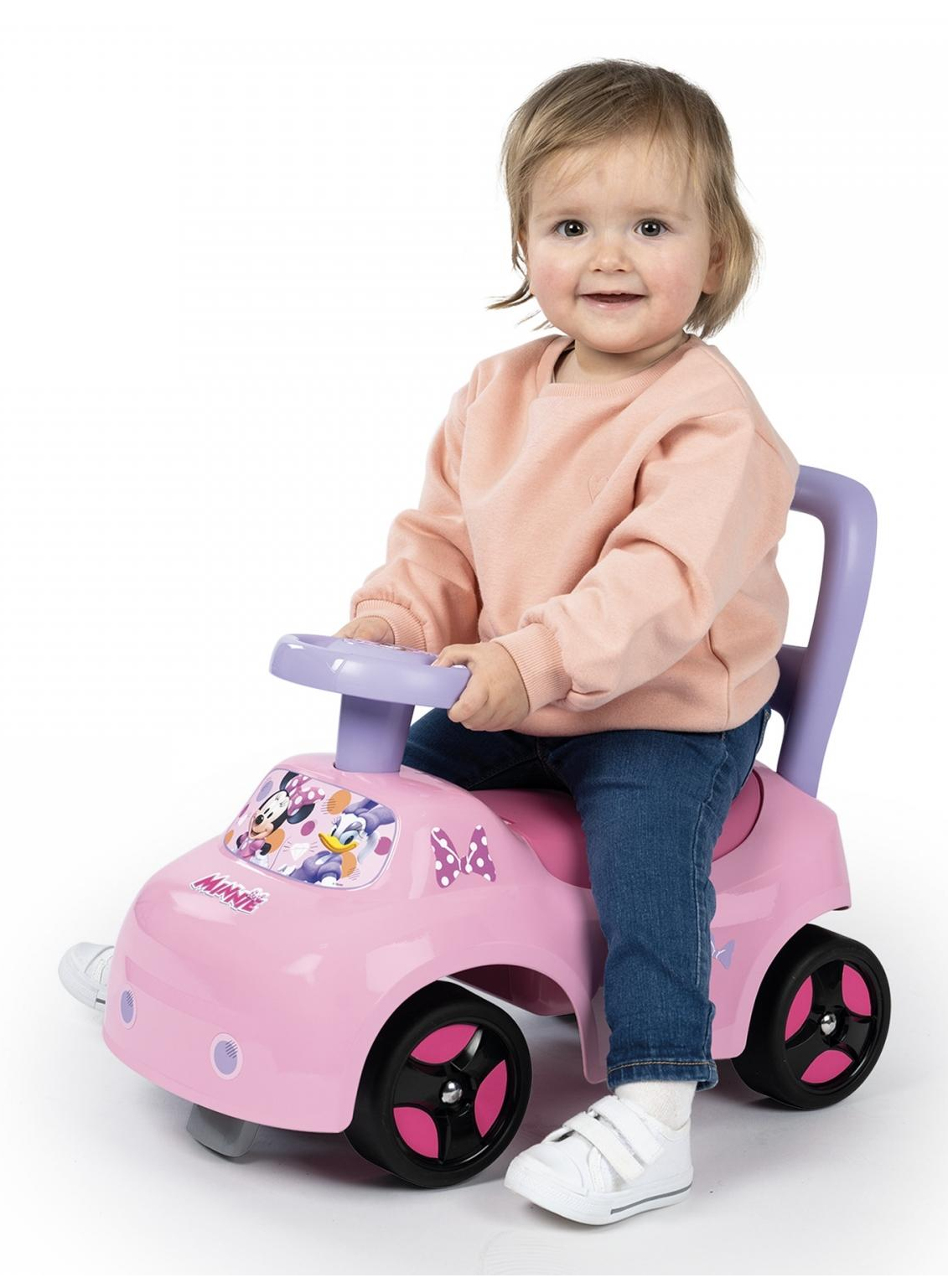 Ein Baby sitzt in einem rosa Spielzeugauto. Sie trägt einen rosa Pullover und Jeans. Weißer Hintergrund.