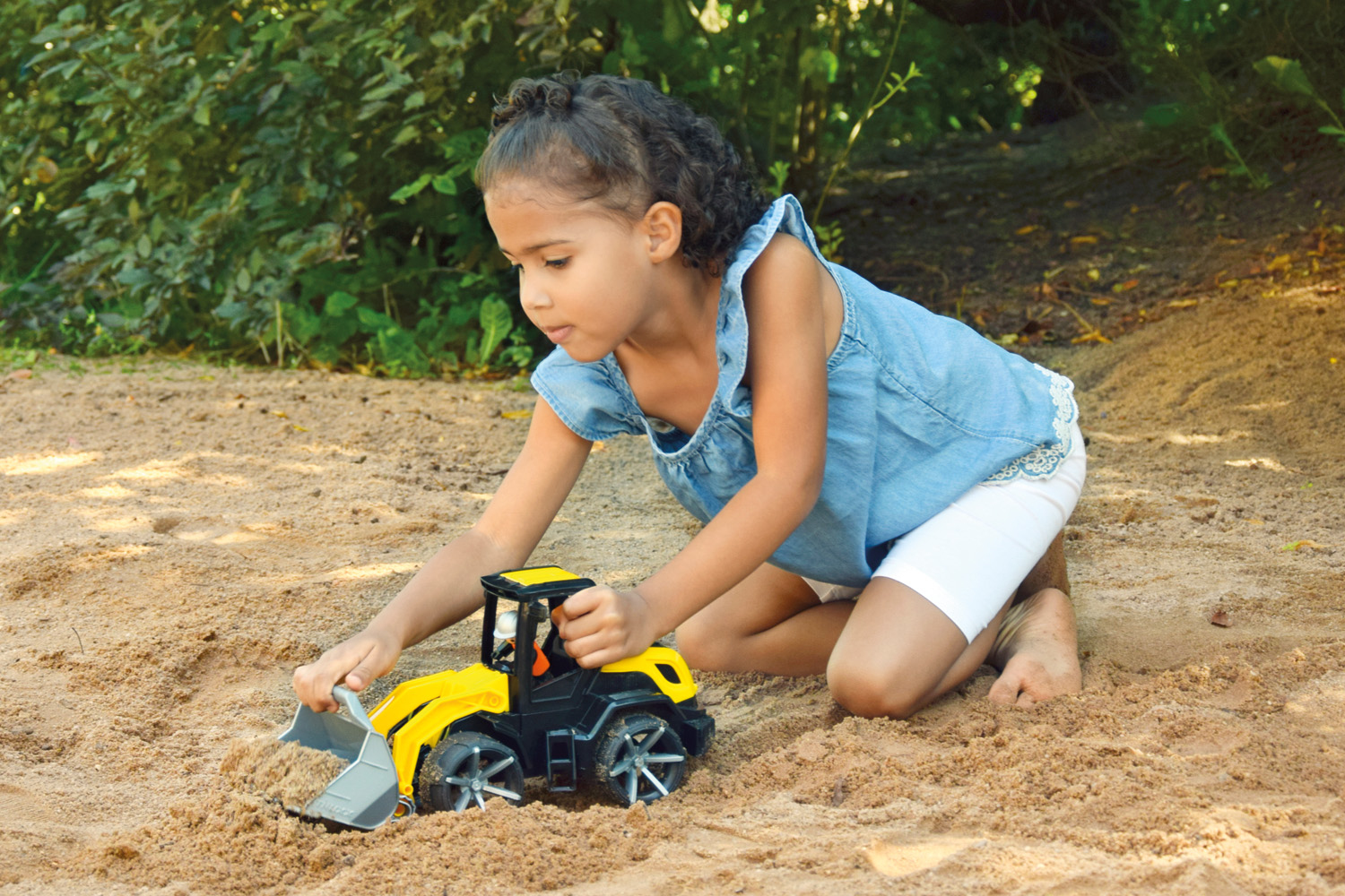 Młoda dziewczyna bawi się żółto-czarną zabawką koparką w piasku. Zielone krzewy w tle.