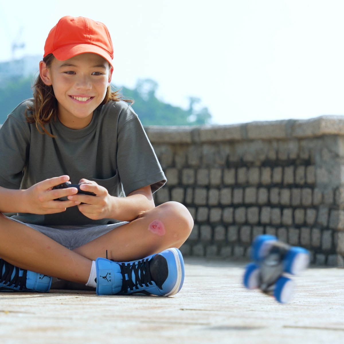 Chłopiec z pomarańczową czapką uśmiecha się, trzyma kontroler, zabawkowy samochód w tle.